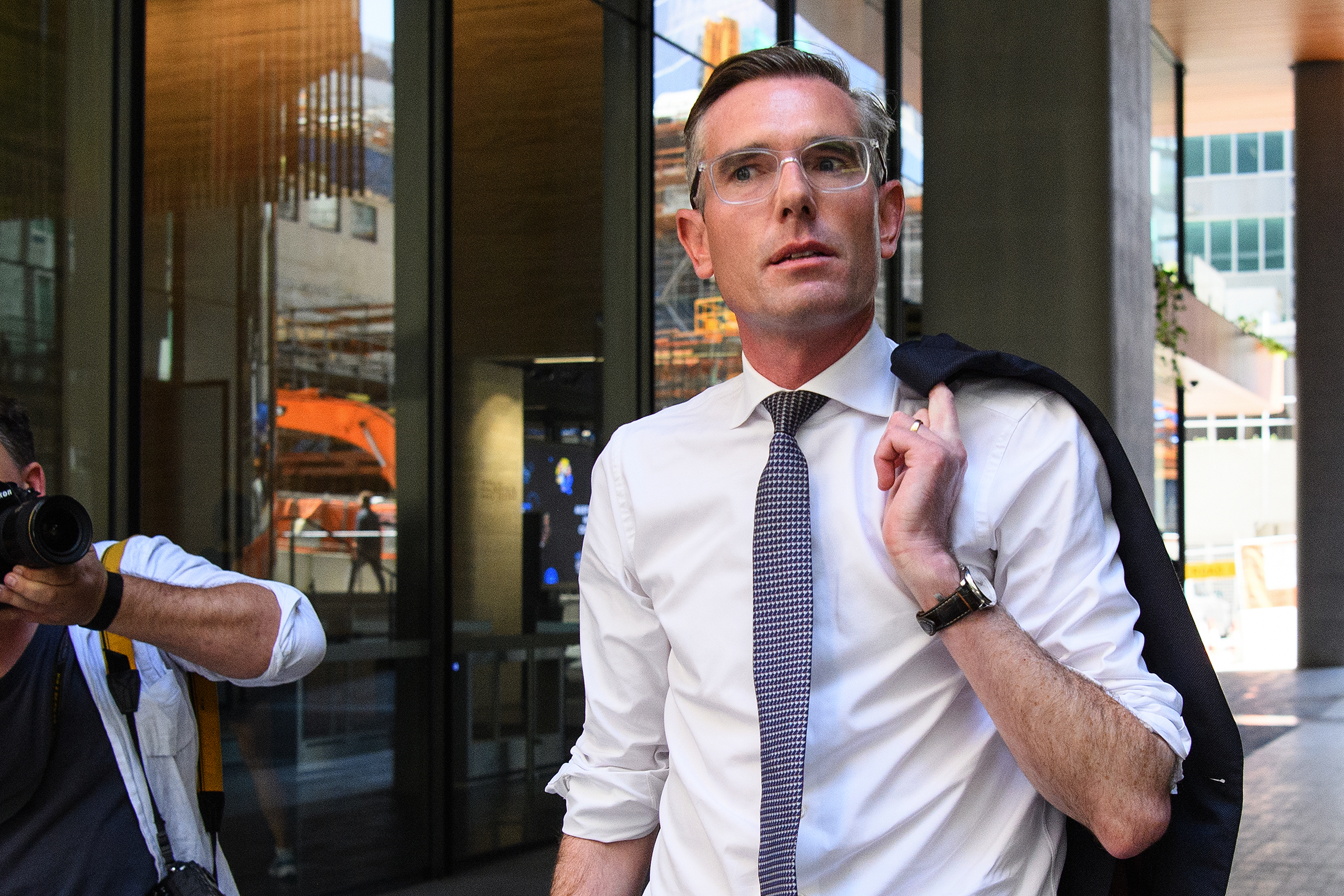 NSW Premier Dominic Perrottet walking down a street in a white shirt and tie. He carries a blazer over his shoulder.