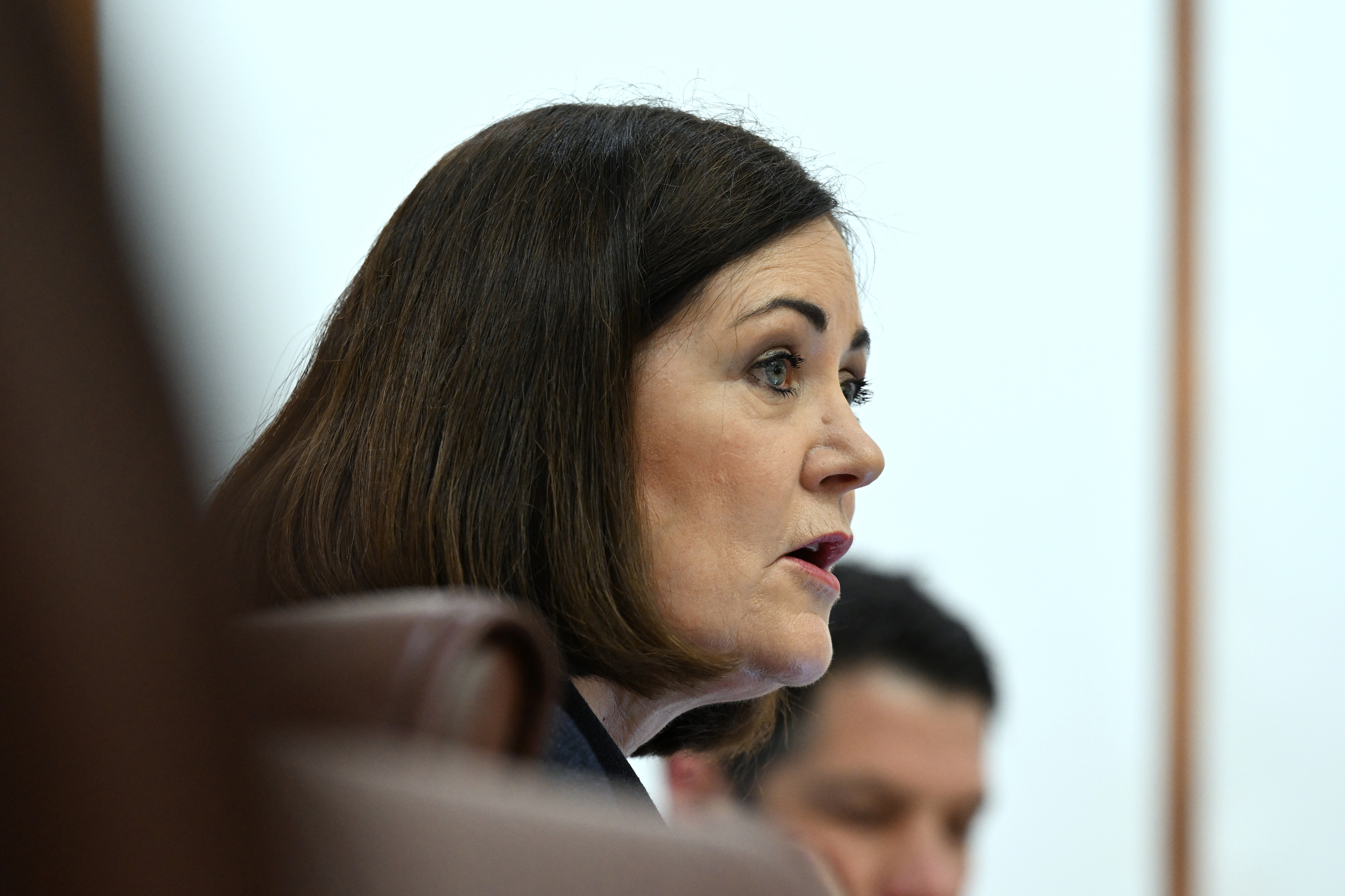 A Caucasian woman with a short blunt brunette bob is shown in a side profile. She is wearing some eye make up and lipstick, and appears to be speaking to someone out of frame.