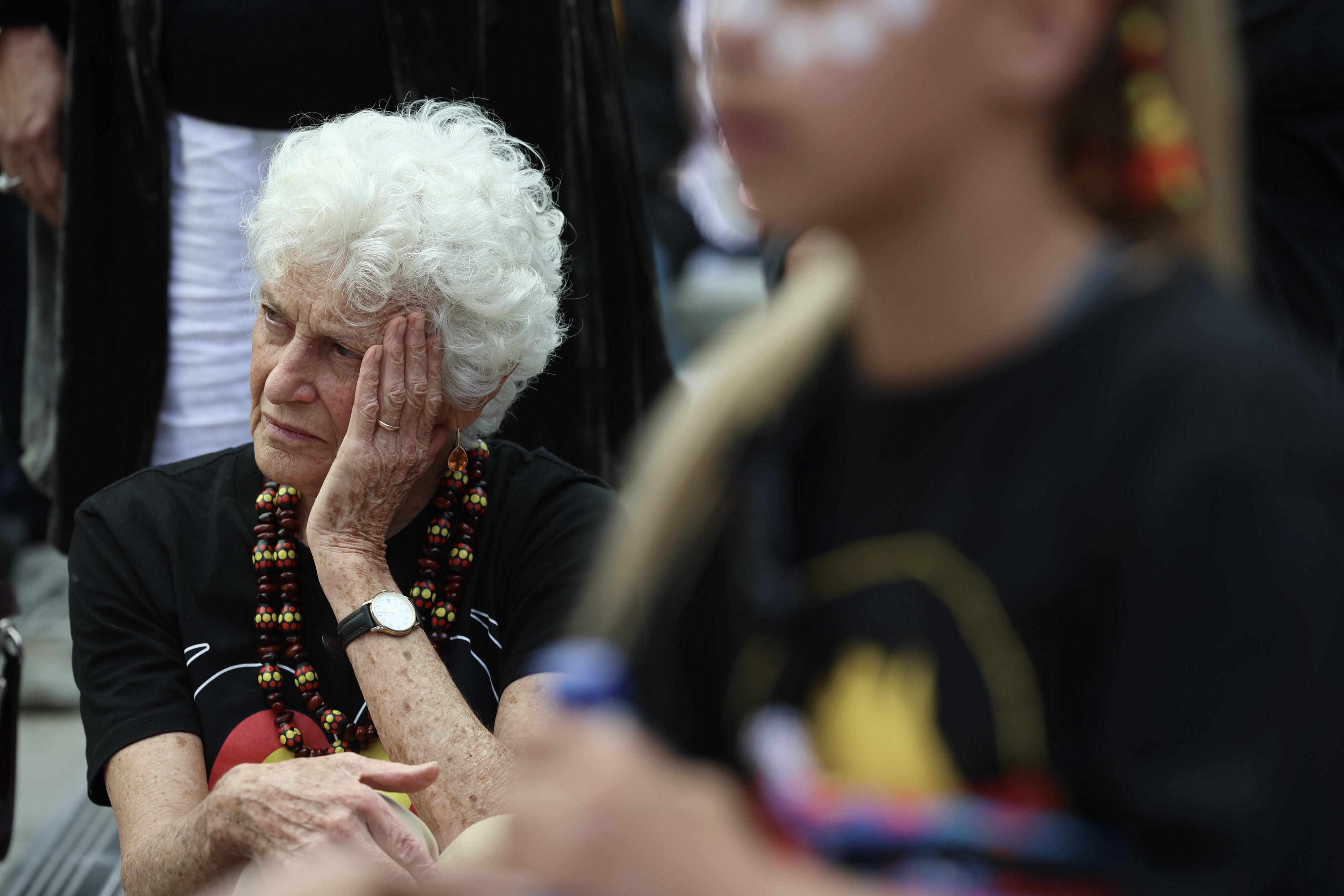 An closeup of an older woman with white hair surrounded by people at a rally. She is seated, holding one of her hands to her head with a sorrowful and tired look on her face. She is wearing a black t-shirt with the Aboriginal colours of red, yellow and black.