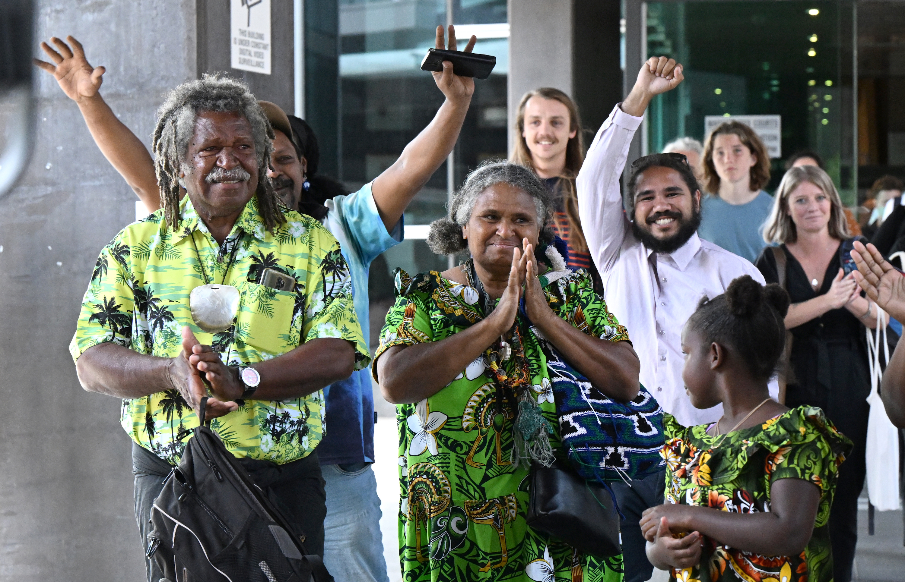 A group of people leaving a courthouse are visibly emotional, with the front three people being a Tiwi Islander family all wearing bright green tropical shirts. There is an older man with grey dreadlocks, an older woman with her hands held in the prayer position, and a young girl looking back towards the woman. 
