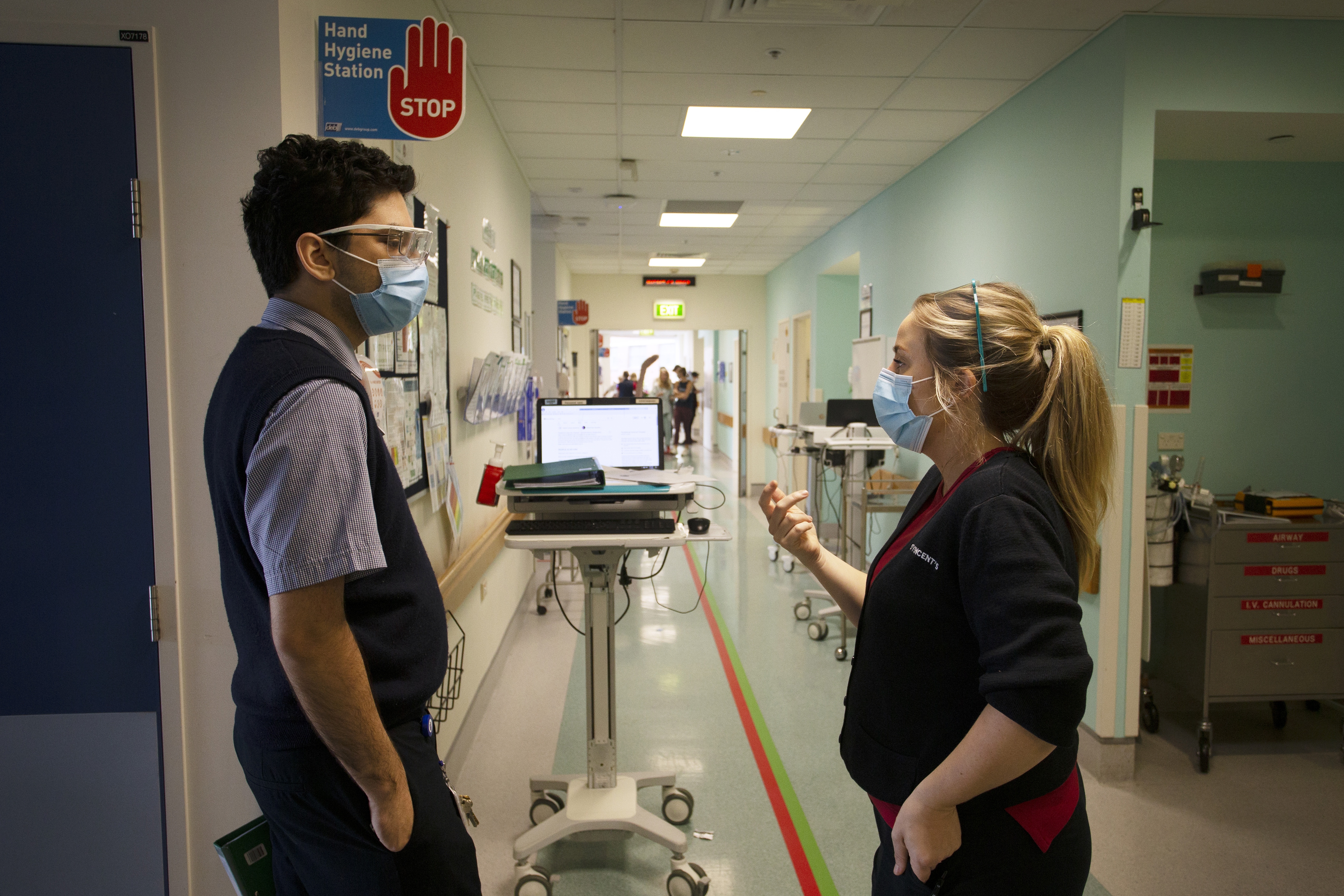 Two healthcare workers in a hospital hallway, facing each other with masks on and in discussion