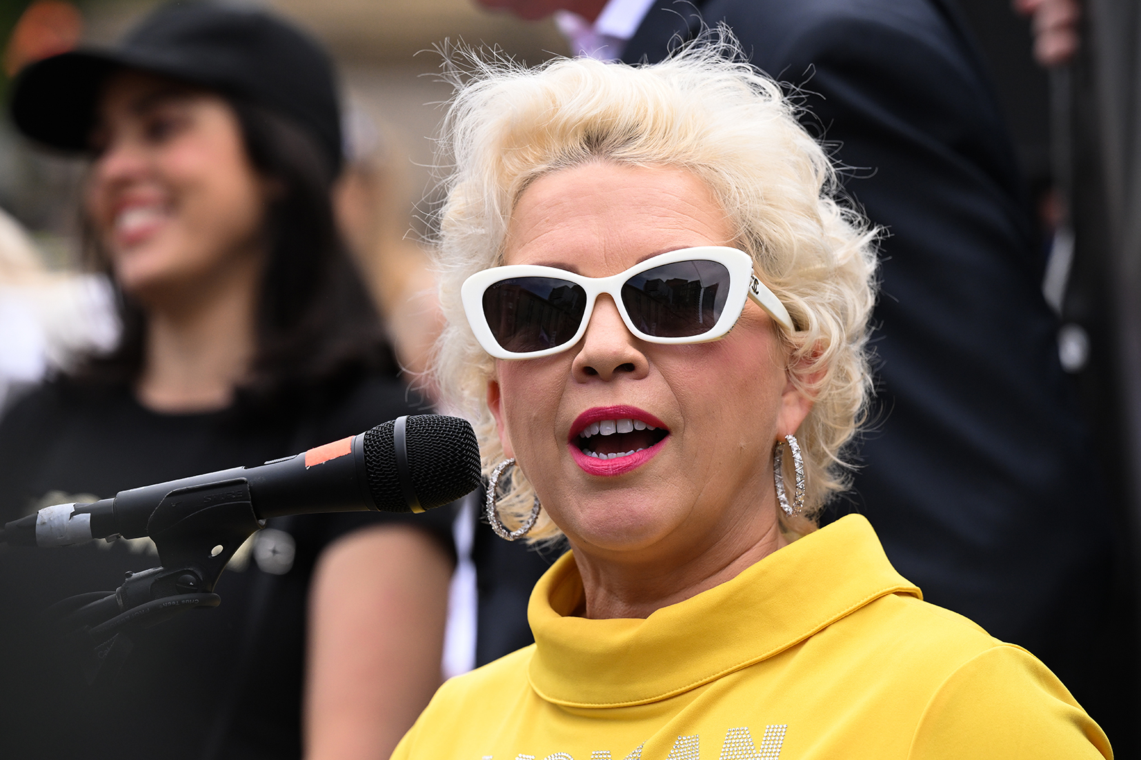 Kellie-Jay Keen-Minshull standing by a microphone and dressed in a yellow top and white sunglasses. 