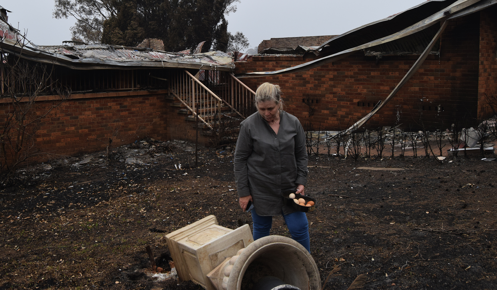 A woman looks down at a fallen statue in the centre of a backyard stripped and blackened by fire.