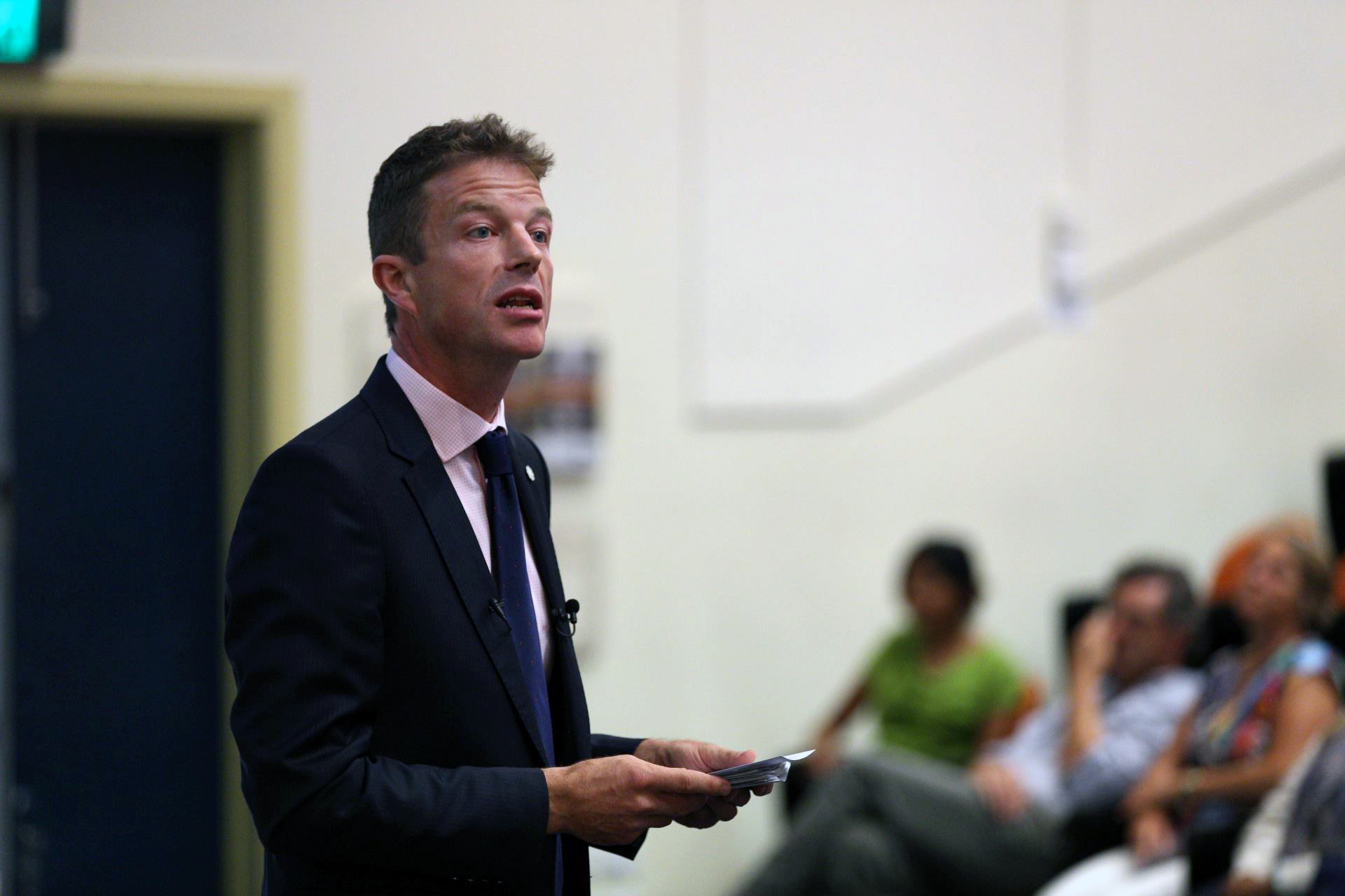 A Caucasian man in his late 30s to 40s in a dark blue suit, pink shirt and blue tie addresses what appears to be a lecture theatre; he is holding note cards