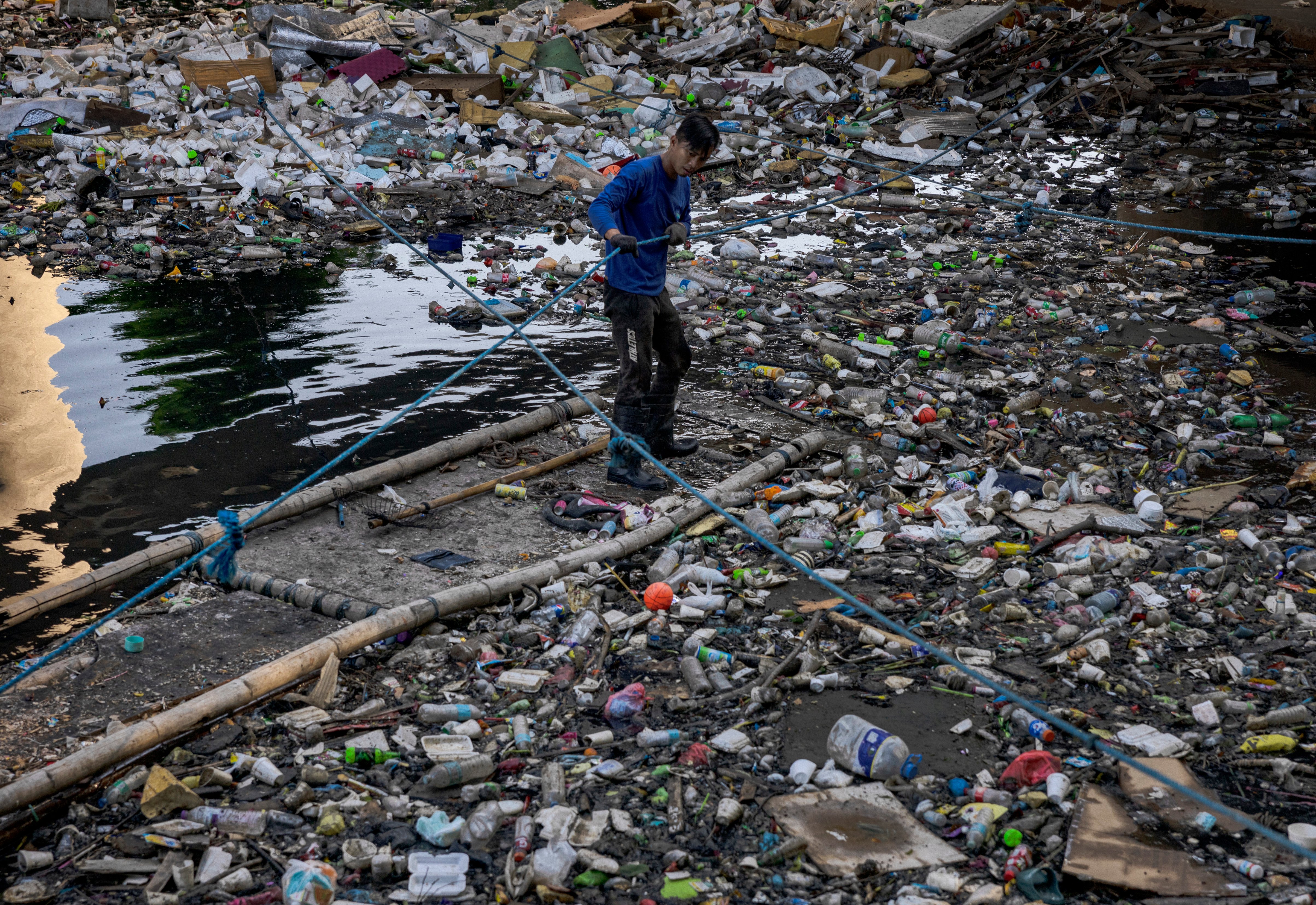 A man collects rubbish by a river filled with plastic.