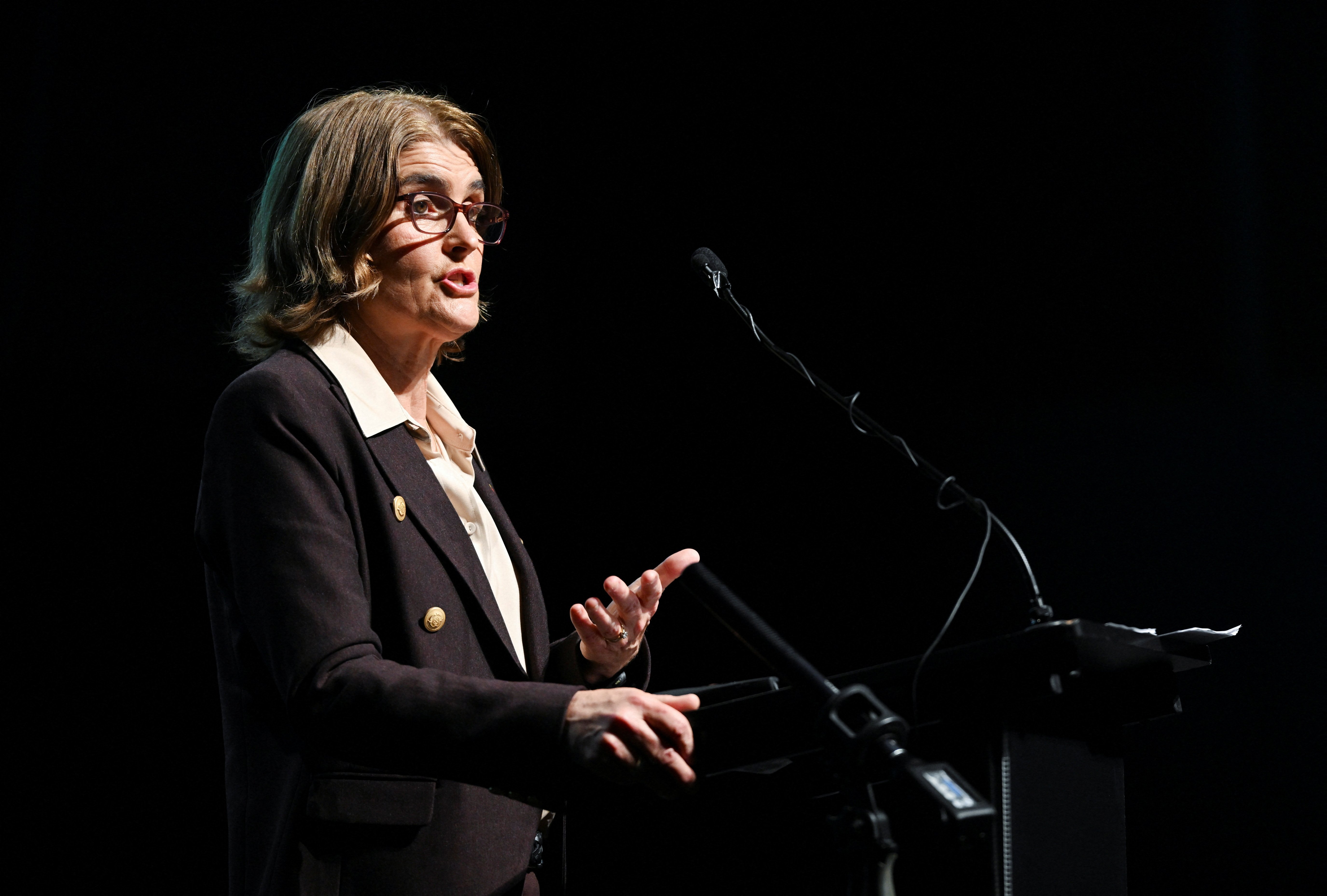 A woman in glasses and a navy blazer speaks at a confeence