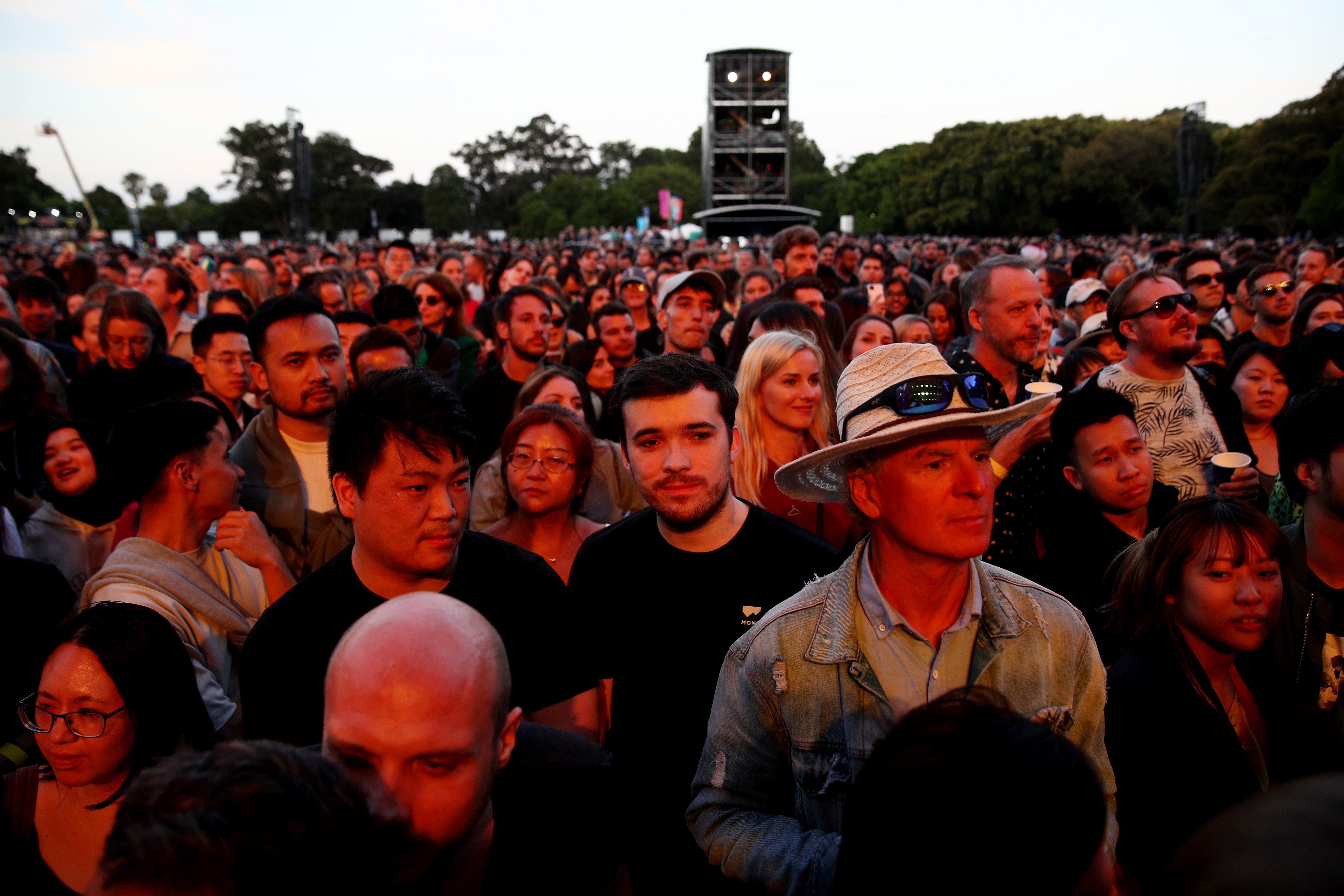 A photo of a crowd watching a gig at sunset, with the lighting red and all people's faces attentive towards the front right of the photo.