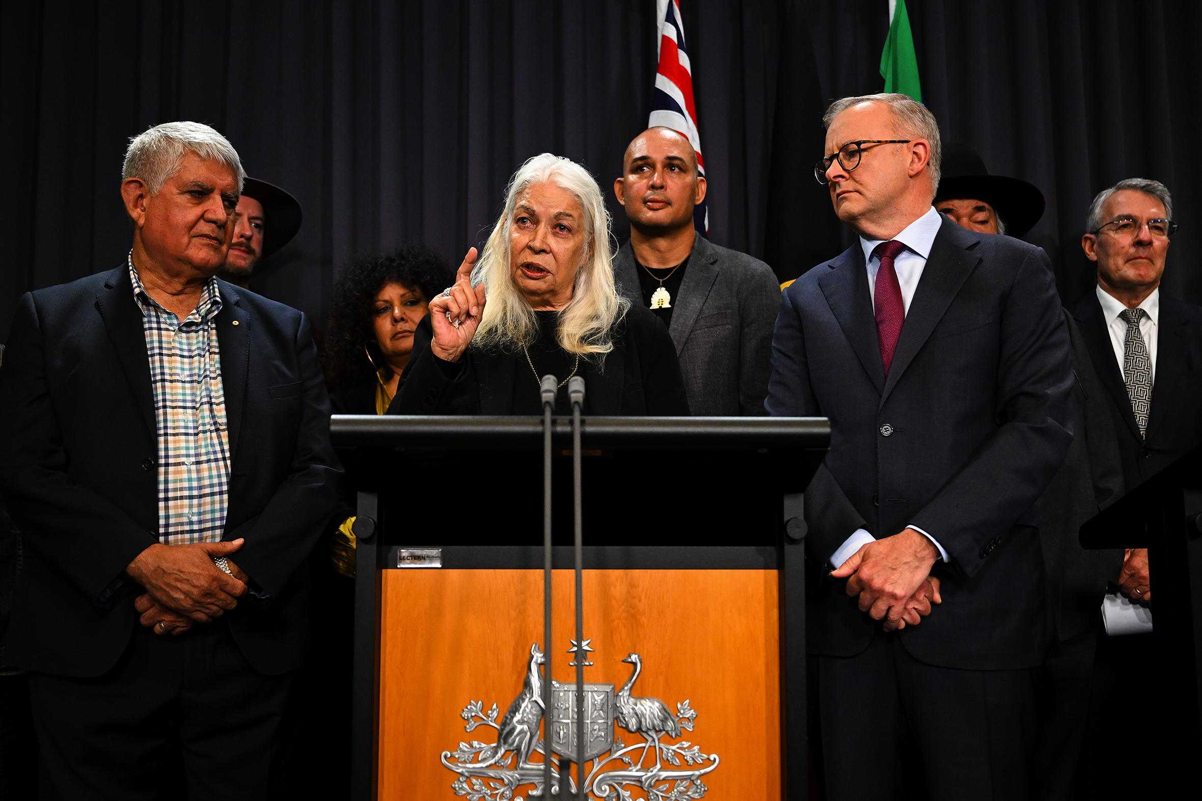A woman with long grey hair speaks at a press conference, flanked by supporters and with the Australian flag in the background.