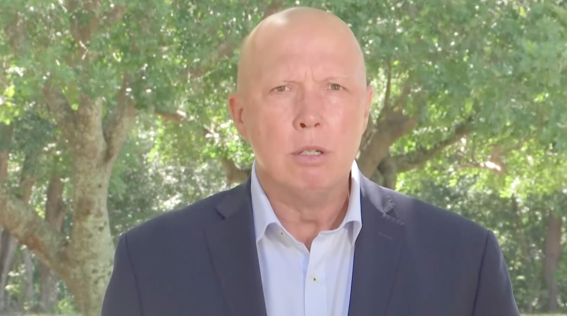 A bald middle-aged Caucasian man appears to be speaking at a press conference. His expression is grumpy and his brow is furrowed. He's wearing a blue suit jacket and shirt, with green trees visible in the background.