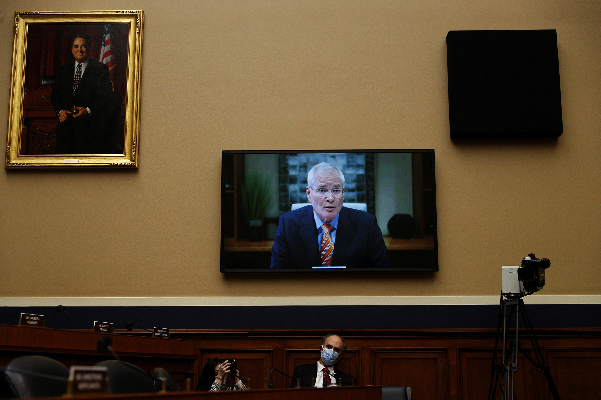 A TV screen in a US Congress building shows a Caucasian man in his 60s with white hair, rectangular glasses and a striped tie who appears to be speaking. Below the TV are two people wearing masks and a television camera.