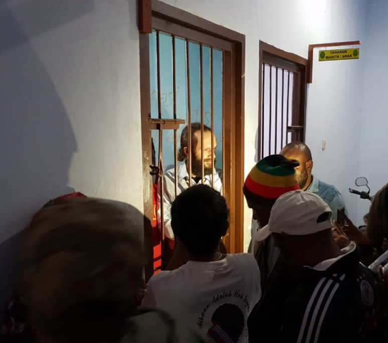 A man with long hair and a beard behind bars. A crowd is gathered around his cell.
