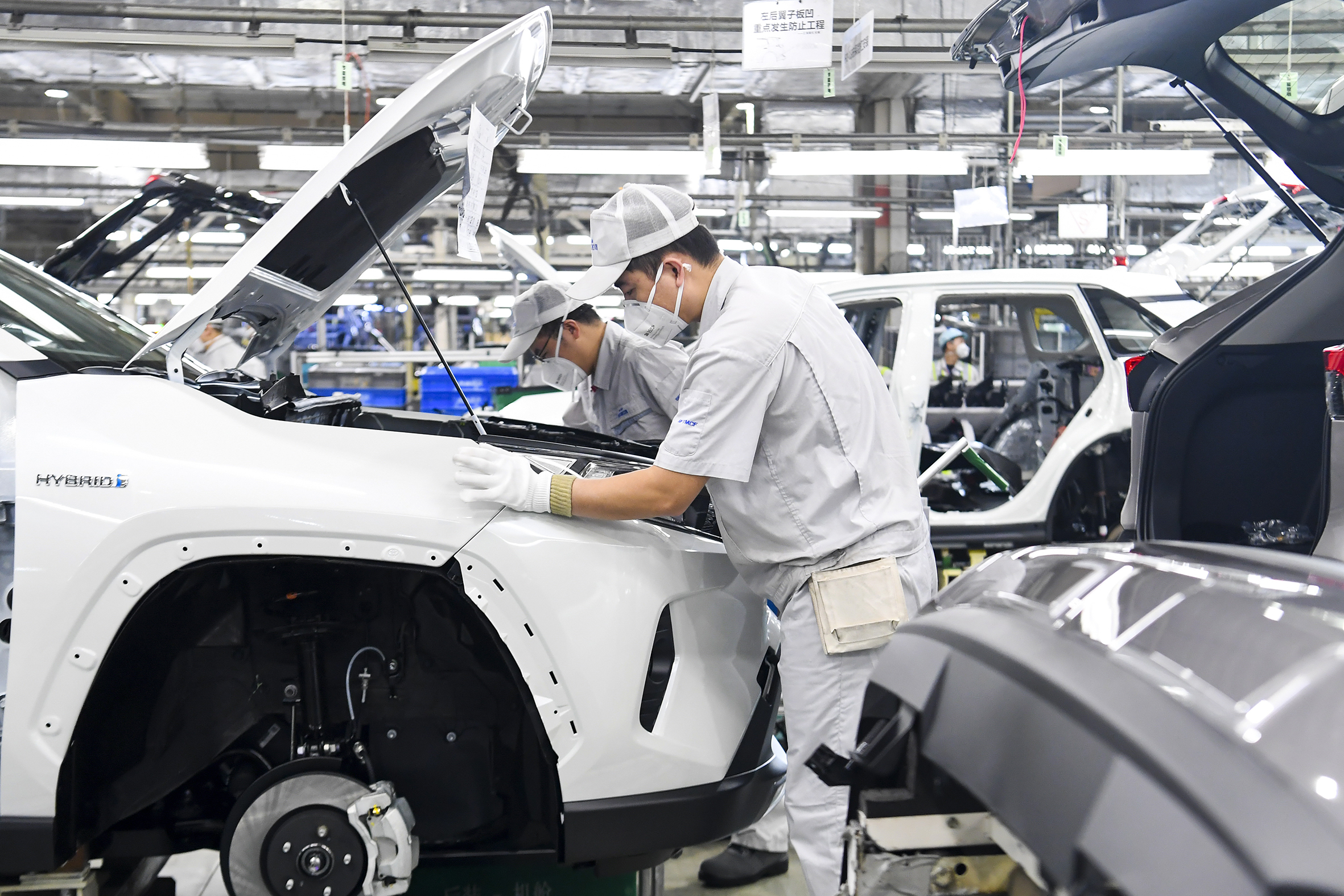 Two workers wearing white clothing, baseball caps and face masks are in a bright car factory surrounded by machinery and parts. The foremost one is looking into the open bonnet of a car.