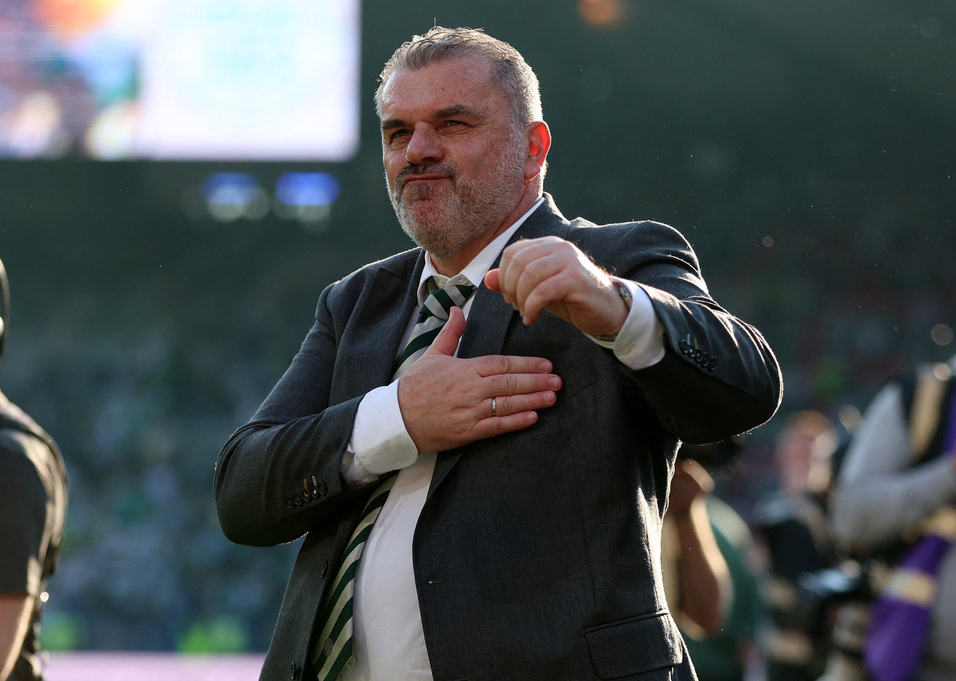 A middle-aged man with grey hair and dressed in a suit claps his hand against his chest, watching on from the sidelines of a football pitch. Behind him is a stadium full of fans.