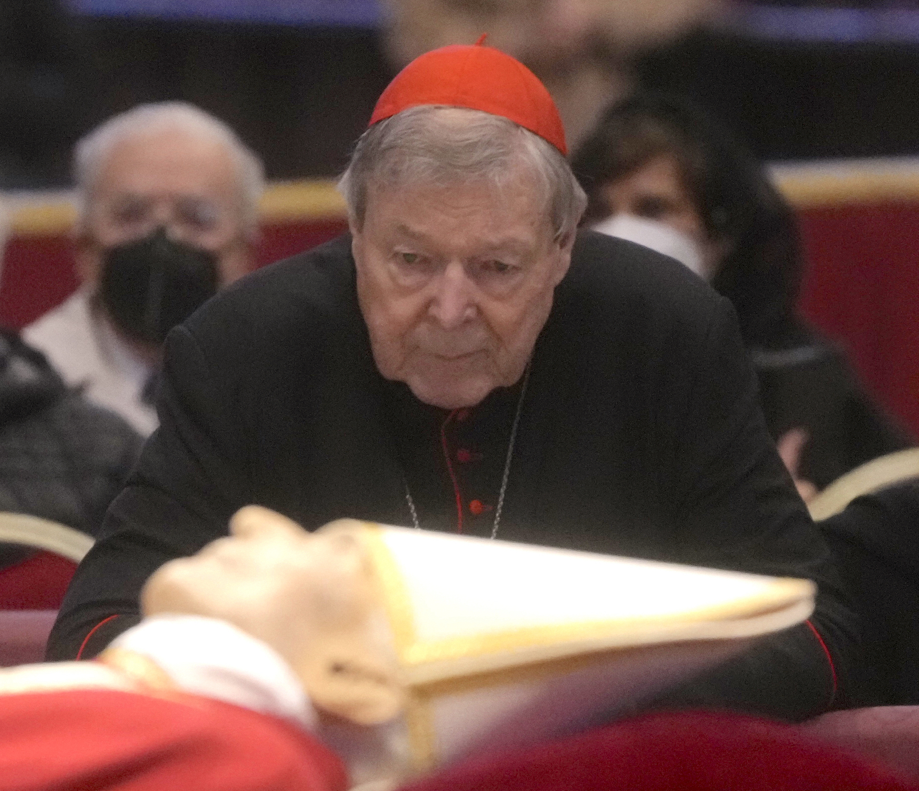 An old Caucasian man with a lined face and saggy jowls wearing a red cardinal's cap and black robes is looking on to the foreground of the picture which is out of focus, but you can see a body dressed in papal robes with a white hat. 