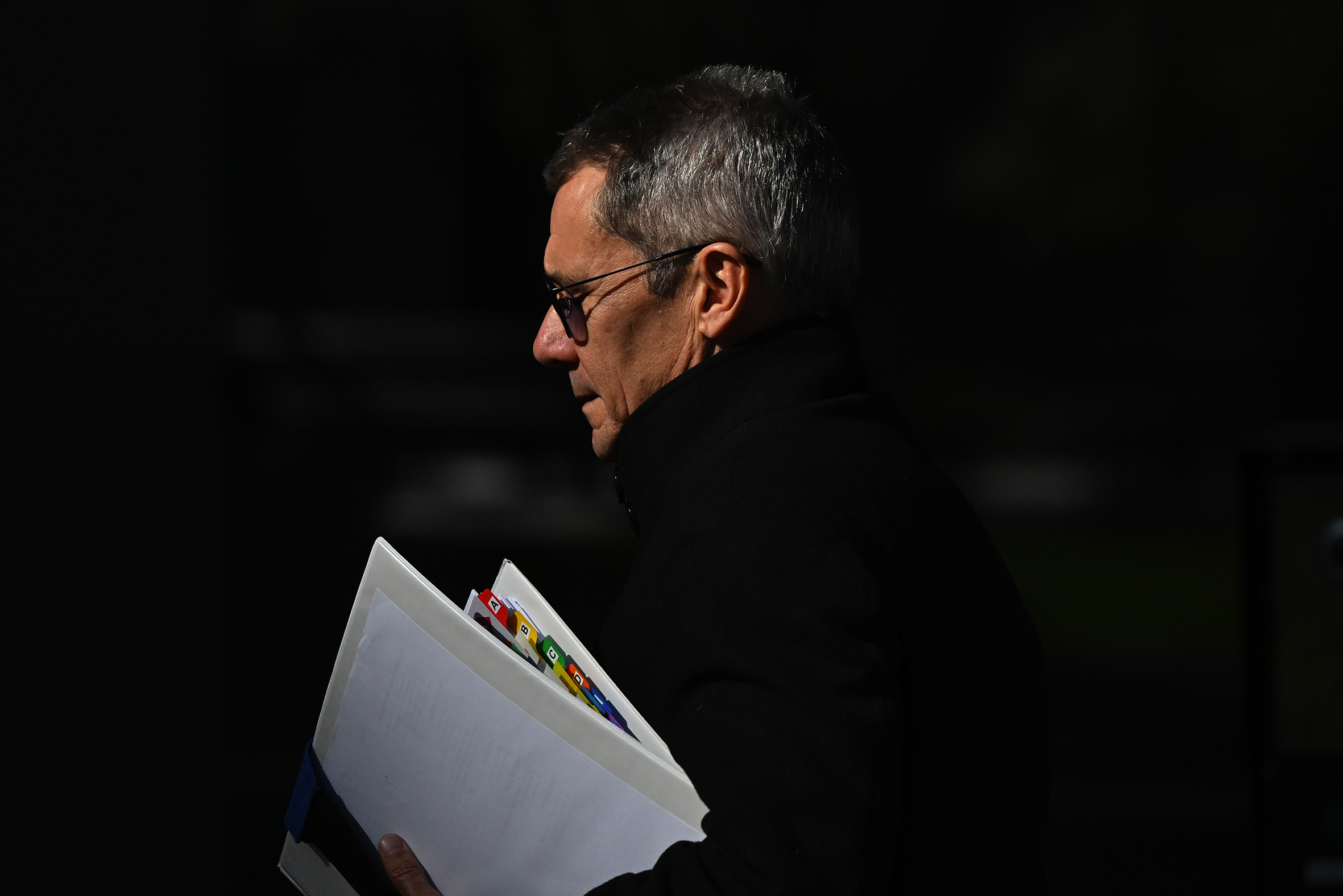 A man wearing a black jacket and glasses walks outside with documents under his arm.