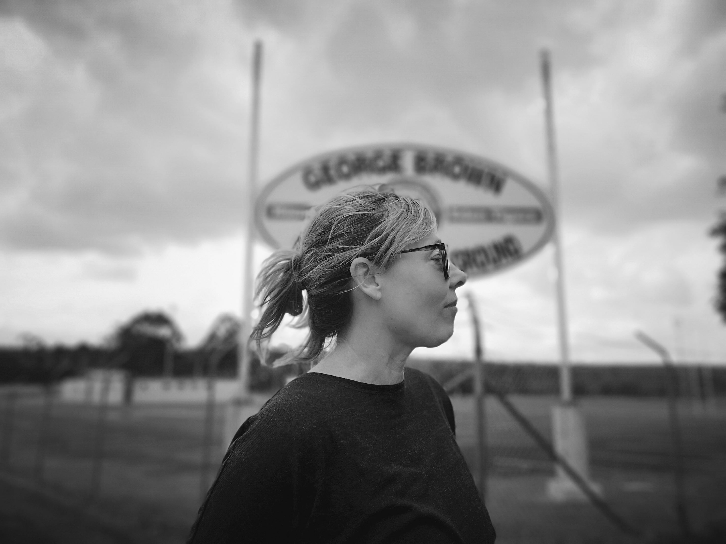 Black and white image of Amanda Brown wearing sunglasses in the foreground, her hair pulled back in a ponytail.