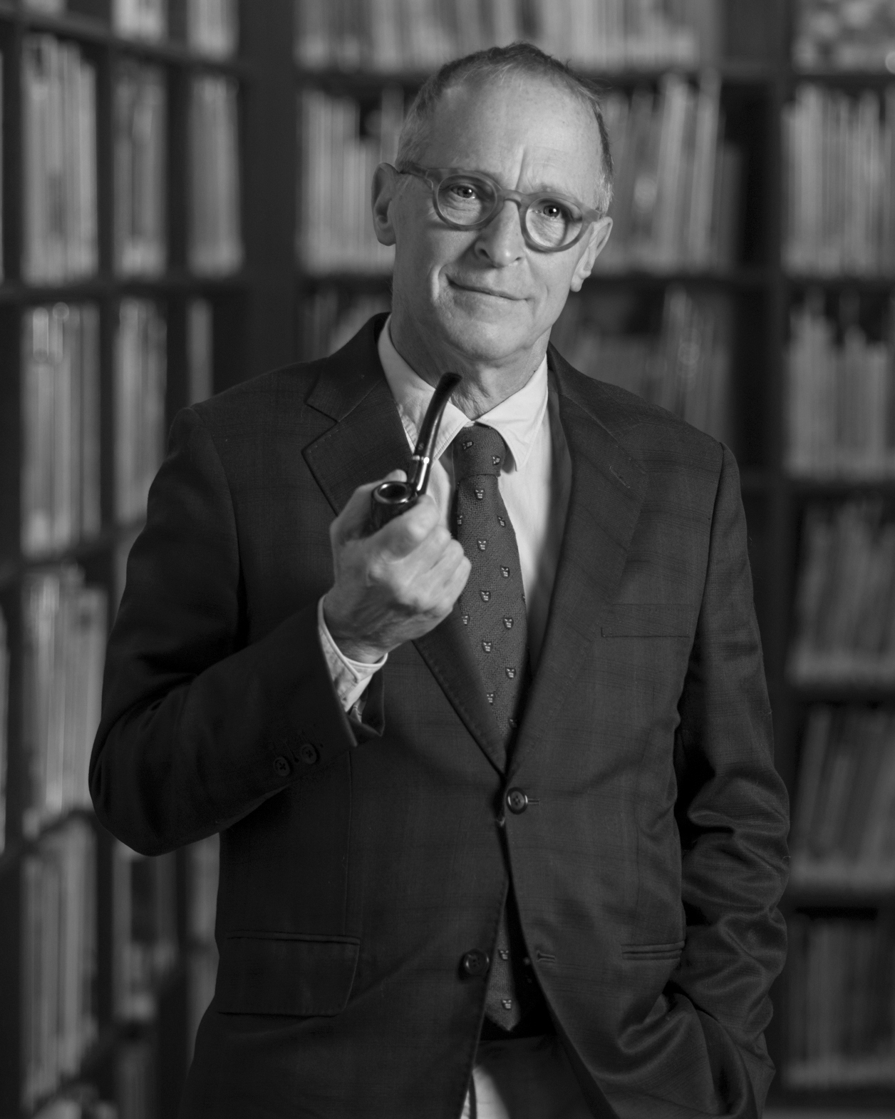 A black and white image of a Caucasian man holding a pipe, wearing glasses and a lovely tailored suit. He has a slight smile on his face, balding hair and kind eyes.