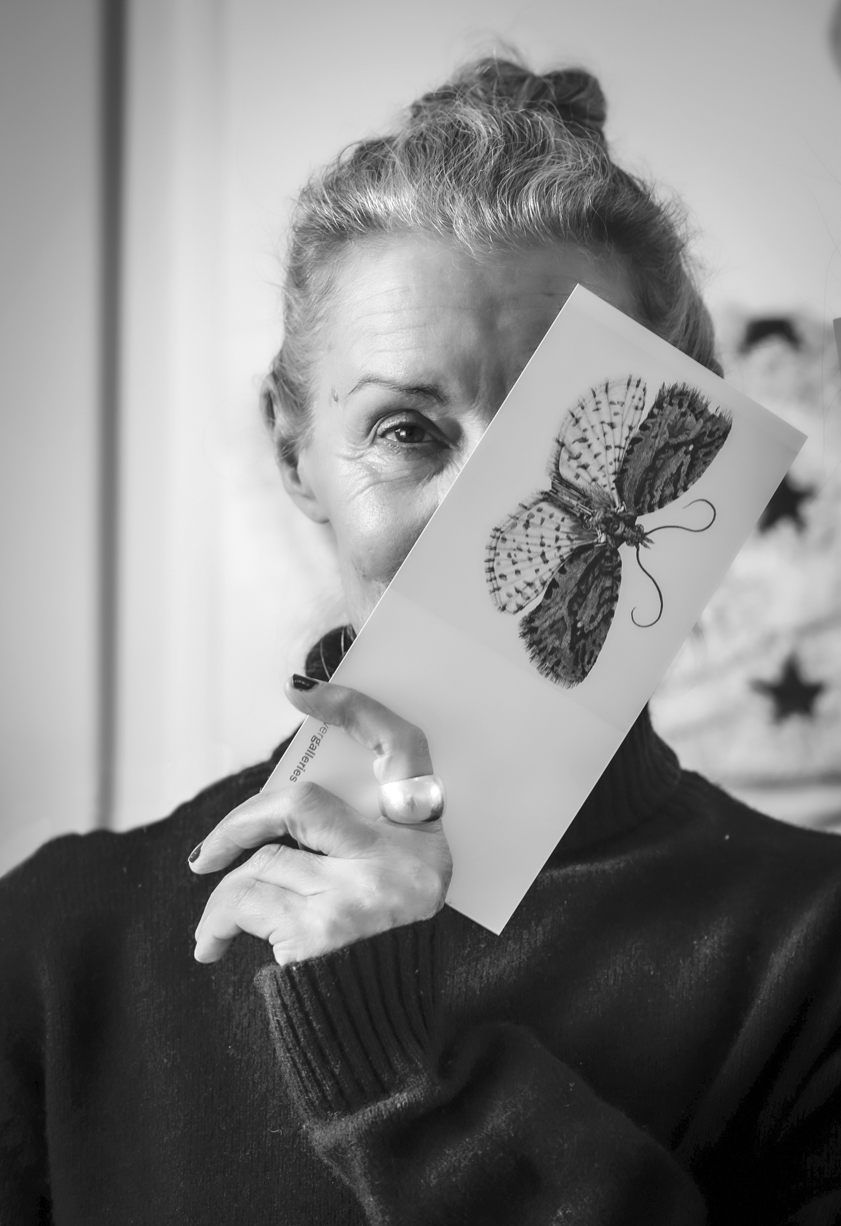 Greyscale portrait photograph of a woman with her hair tied into a bun, revealing only one eye as she obscures her face with a bookmark.