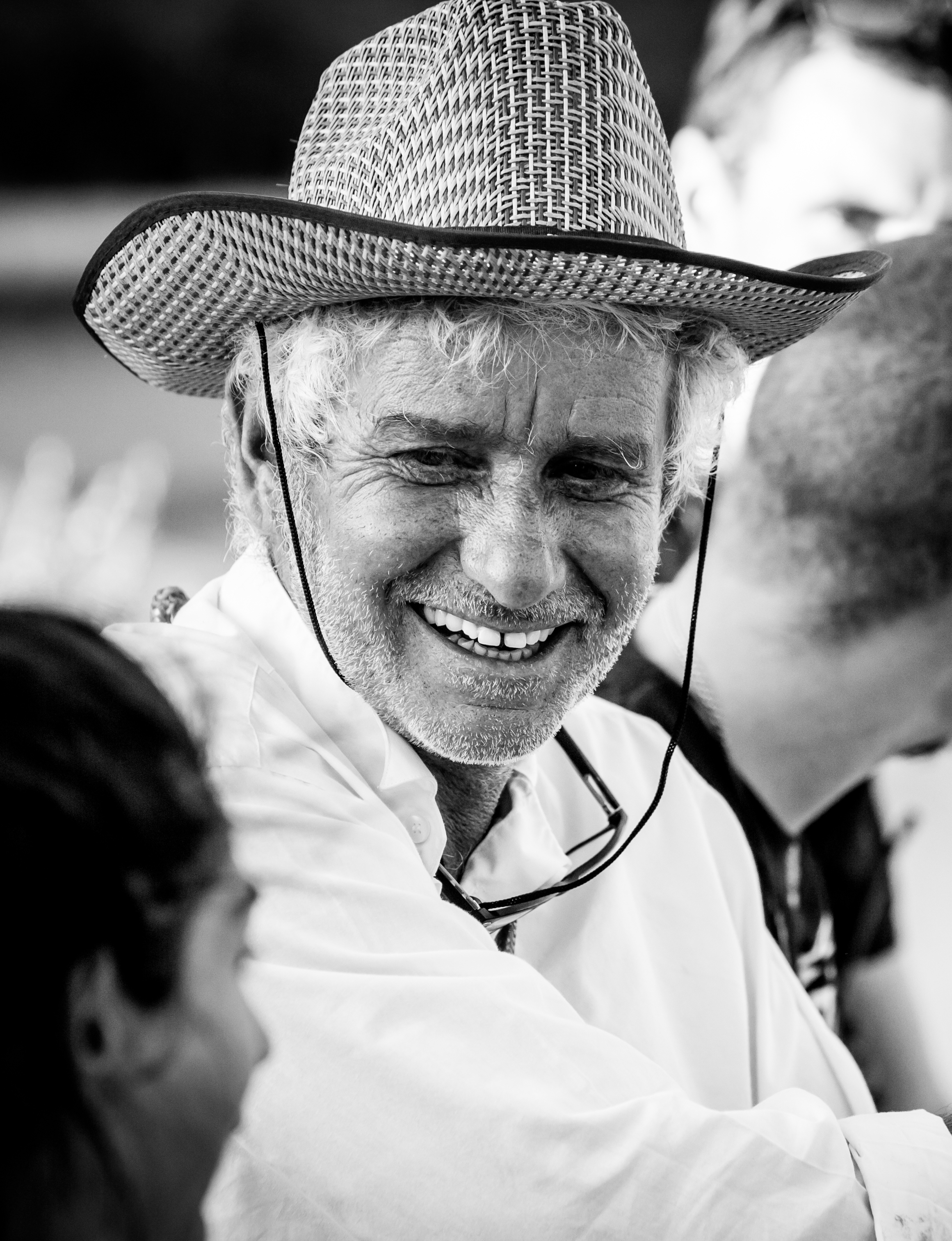 A black and white image of a Caucasian man wearing a straw cowboy hat and a white shirt. His face is grizzled and he has a salt and pepper beard. He is smiling at a woman to his right who is out of focus. 