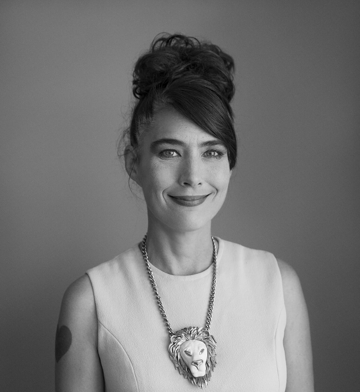 Black and white portrait photograph of Kathleen Hana – a smiling, middle-aged woman wearing her dark hair tied in a bun matched with a necklace that is attached to a lion pendant. A heart is tattooed on her right shoulder.