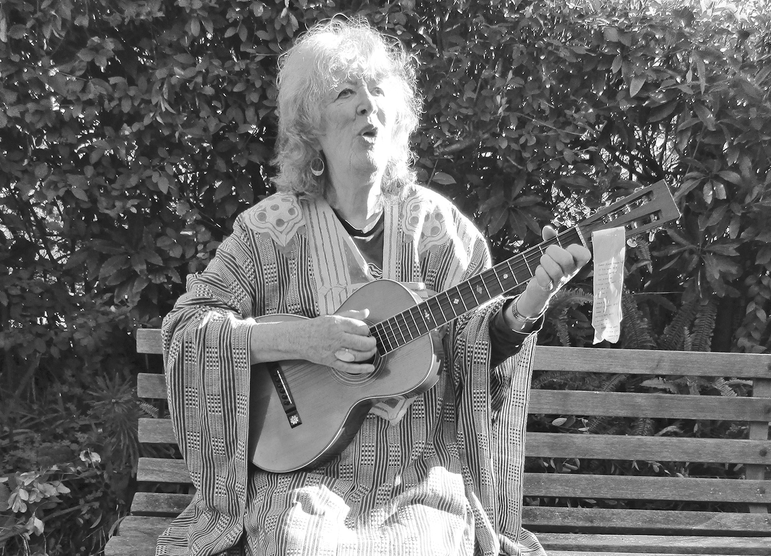 Greyscale image of an elderly woman sitting on a park bench as she plays a guitar.