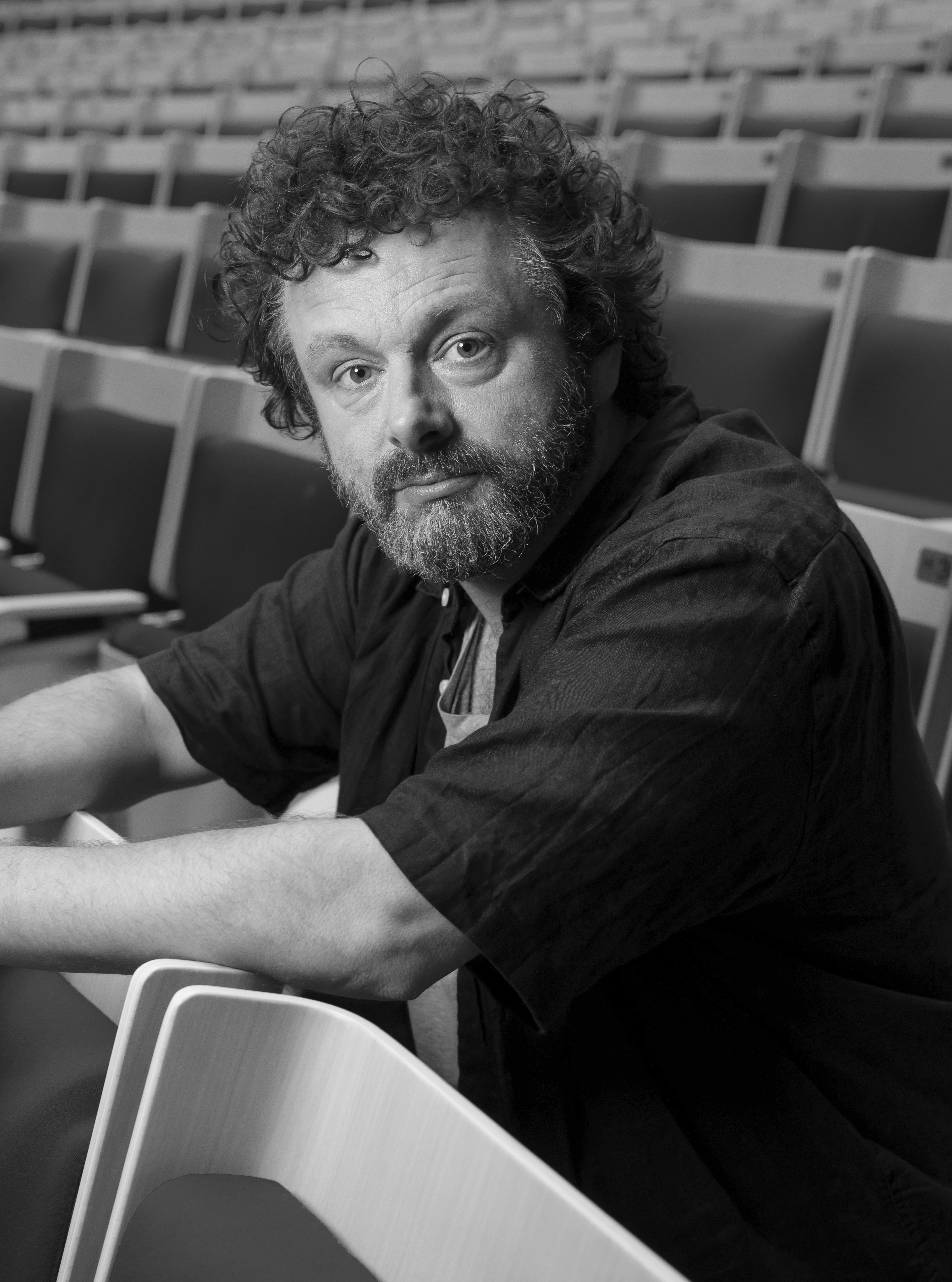 A slightly dishevelled Caucasian man with curly hair and a greying beard sits in an empty theatre