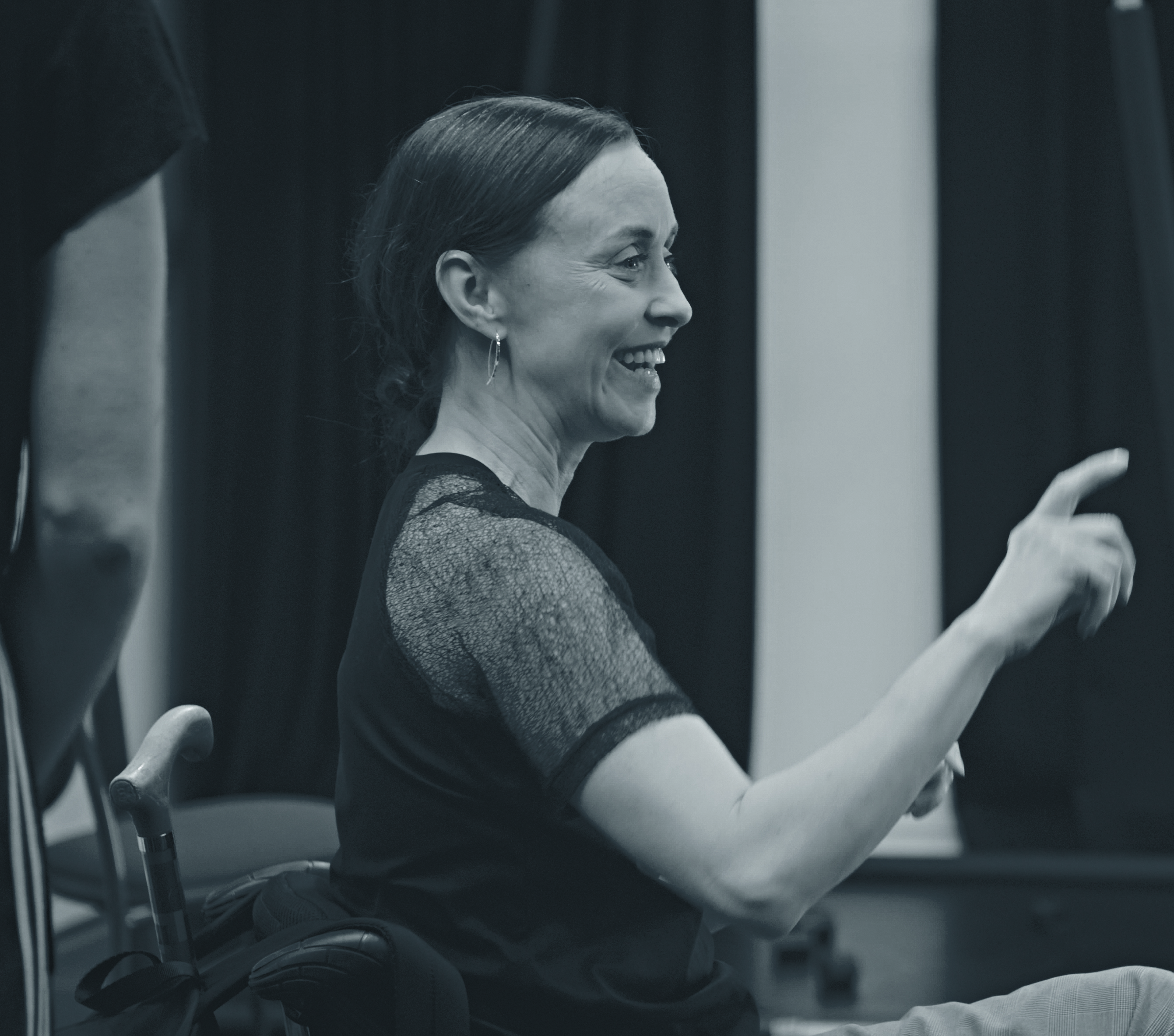 Black & white image of a woman sitting in a wheelchair, smiling with her arm raised.