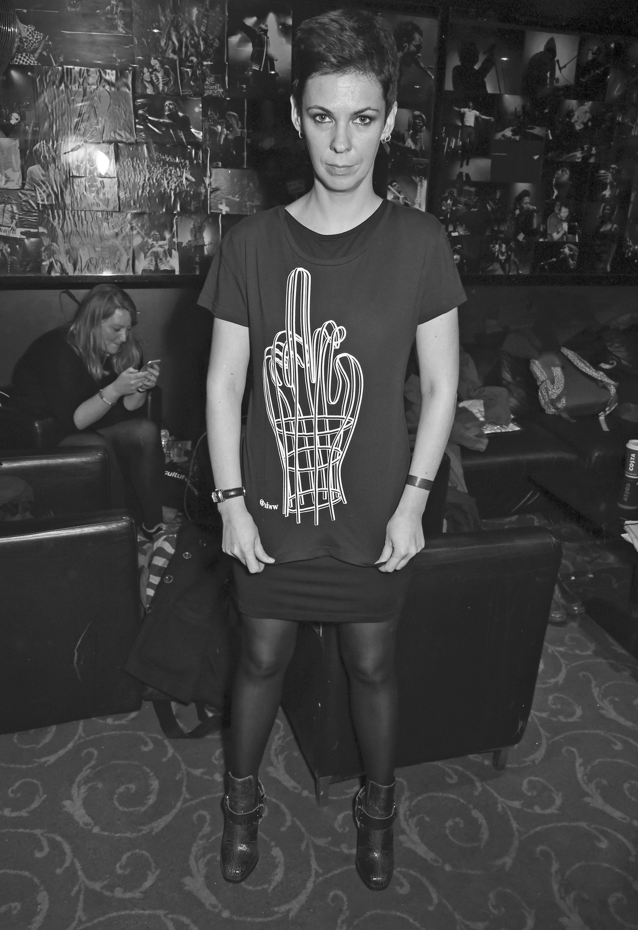 Photograph of Natalia Kaliada standing in a venue with a collage of rock performers covering the wall behind her. She has blue eyes, short hair, and is wearing looped earrings with a t-shirt that has a sketch of a hand with a raised middle finger printed on the front.