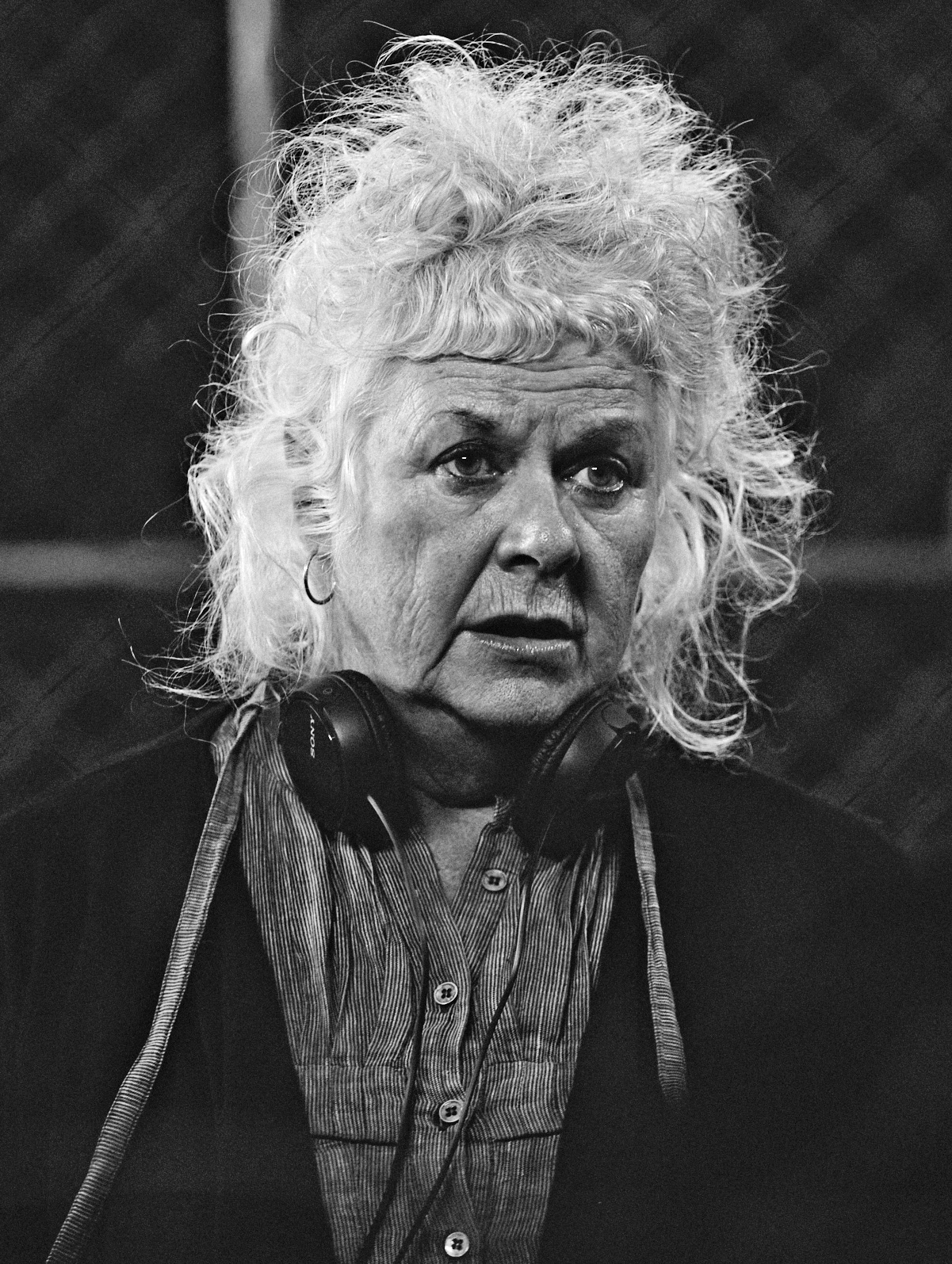 An older Caucasian woman with a mess of white frizzy hair and a short fringe wearing headphones around her neck and a striped shirt looks to the right of the image. Her expression is intense and her face is lined.
