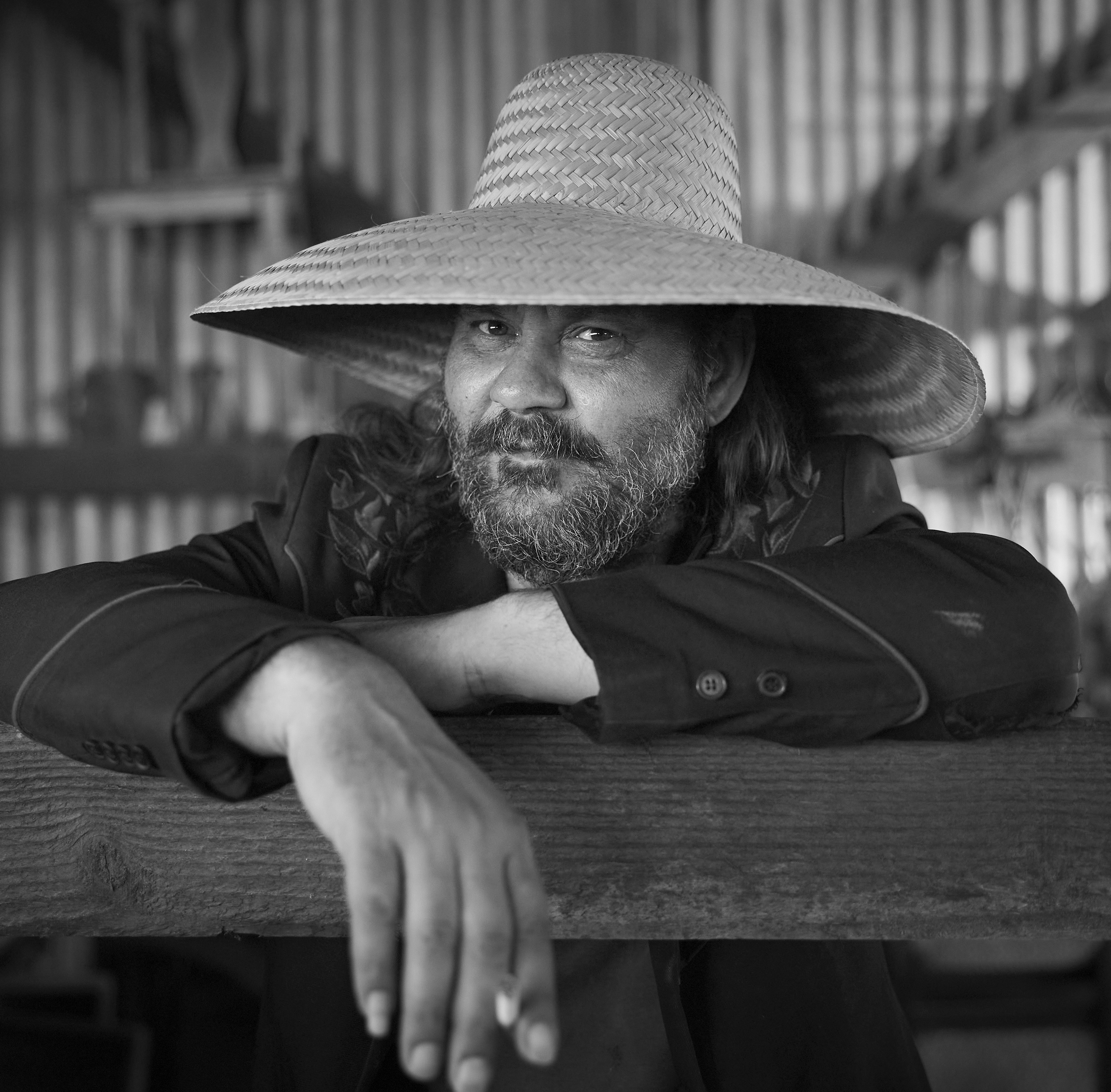 Greyscale image of a middle-aged man with a greying beard wearing a straw hat as he leans against a post.