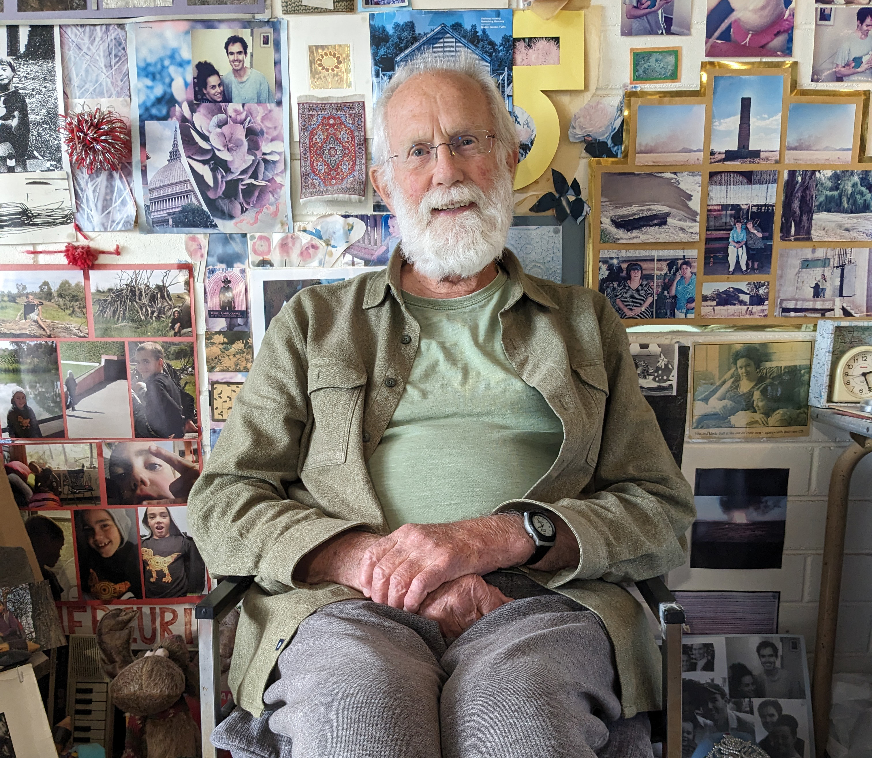 An elderly man surrounded by pictures on his wall.