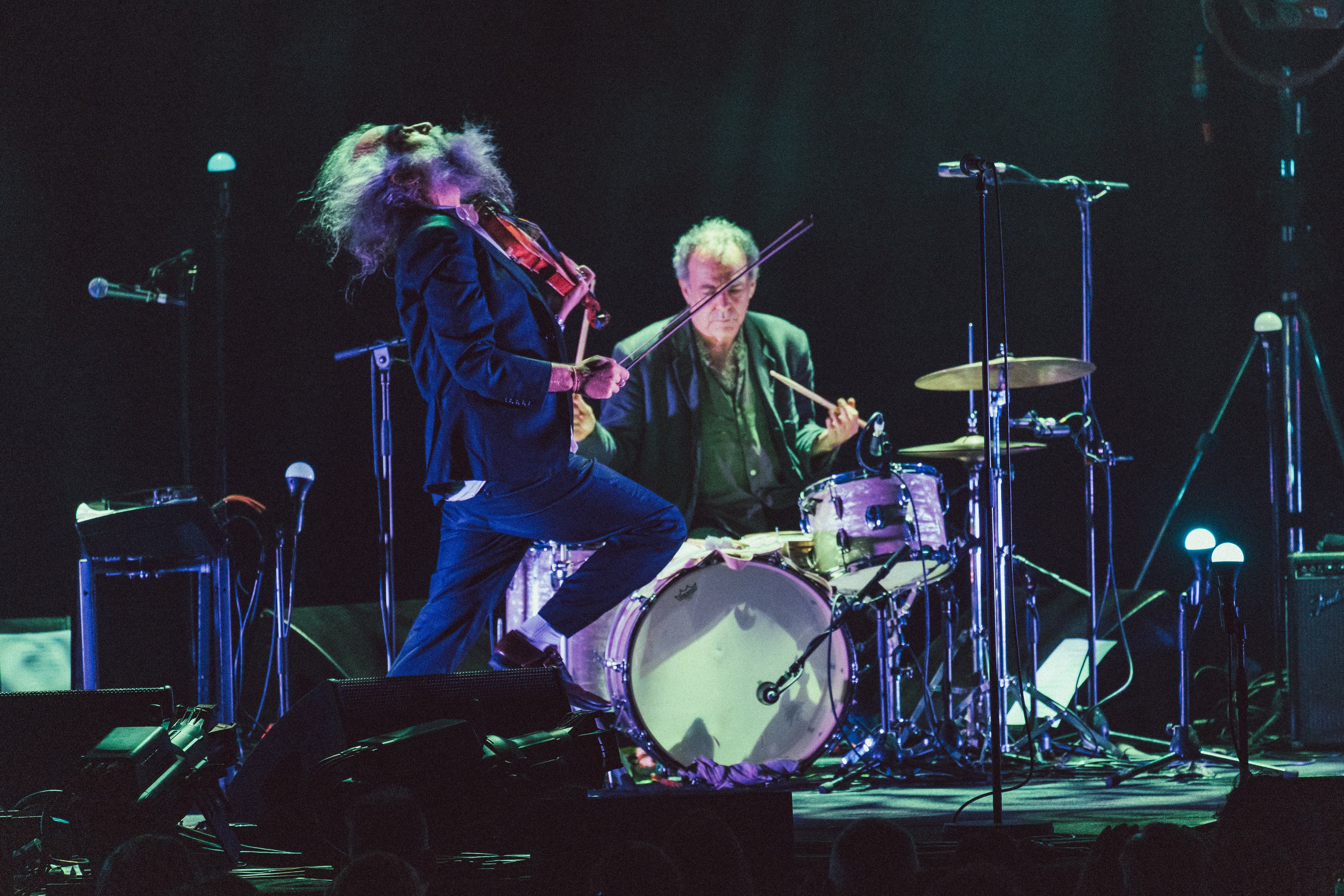 Warren Ellis holds a violin on stage and leans back, his hair unruly. John Ellis sits at his drums mid-beat. 