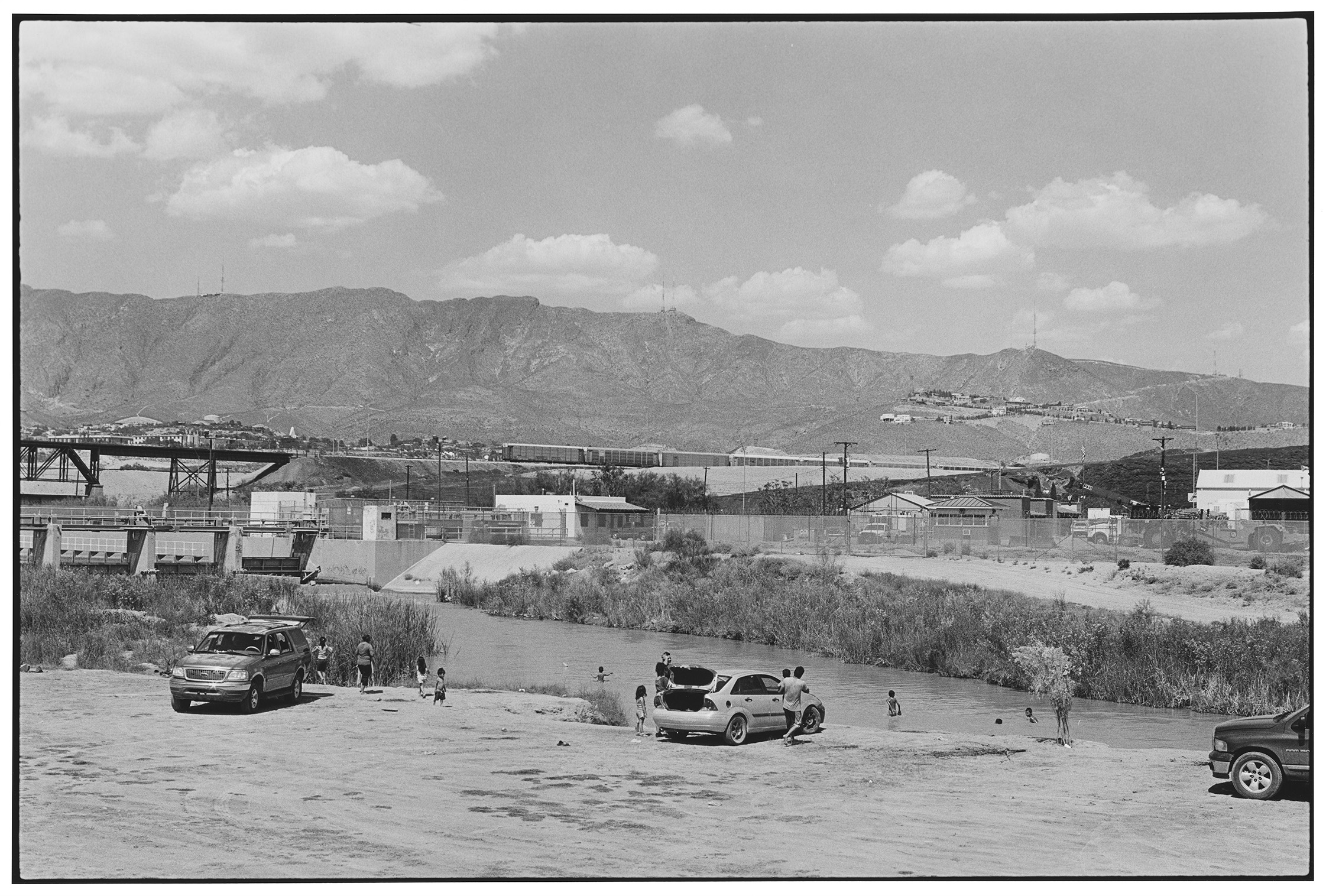 Black & white photograph of families swimming in a river with a fenced country border in the background.