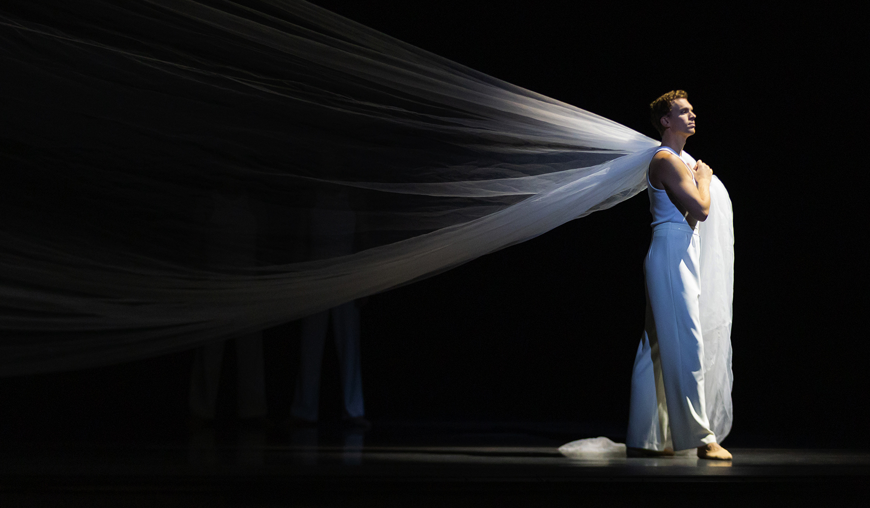 A ballet dancer stands on stage in all white. A hanging white curtain is draped over their shoulder.