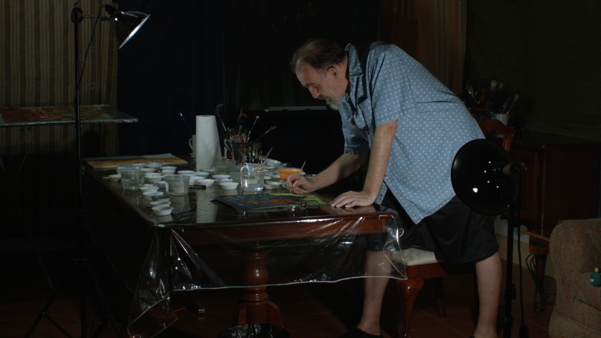 A balding man with a beard paints at a table.