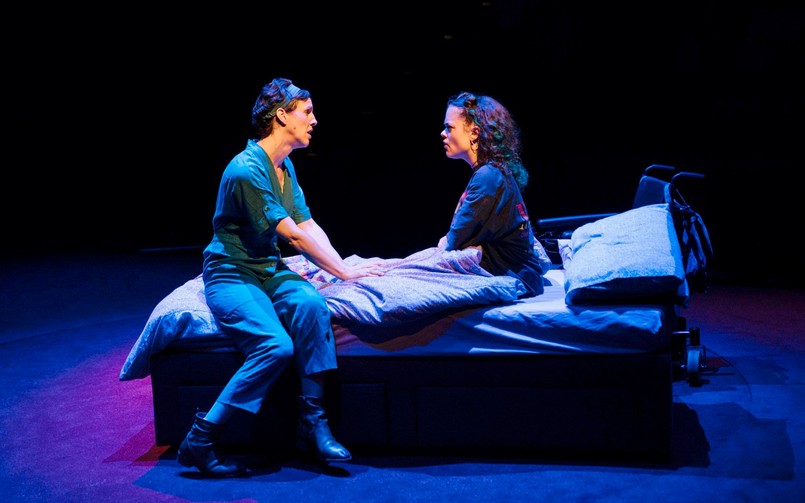 Two women in discussion as they face each other while sitting on a bed. A wheelchair is in the background.