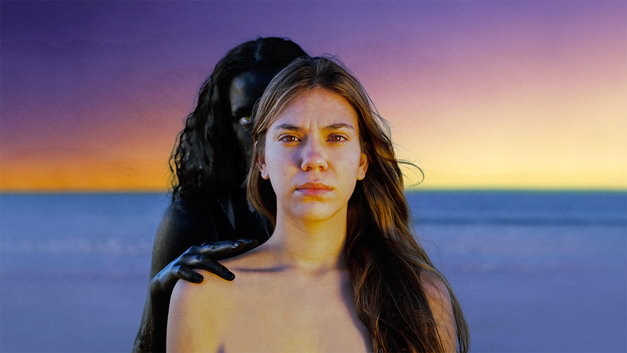 Portrait photograph of a woman with a dark figure behind her.In the background is the ocean and a golden sunset.