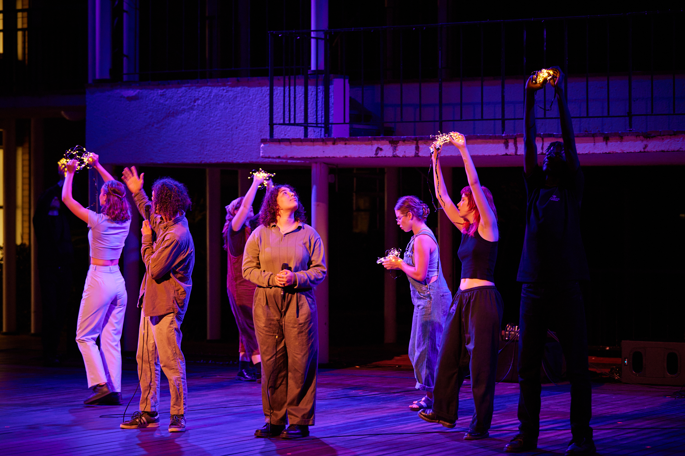 A group of performers on stage. Two hold microphones while several others hold LED lights, which they have raised above their heads.