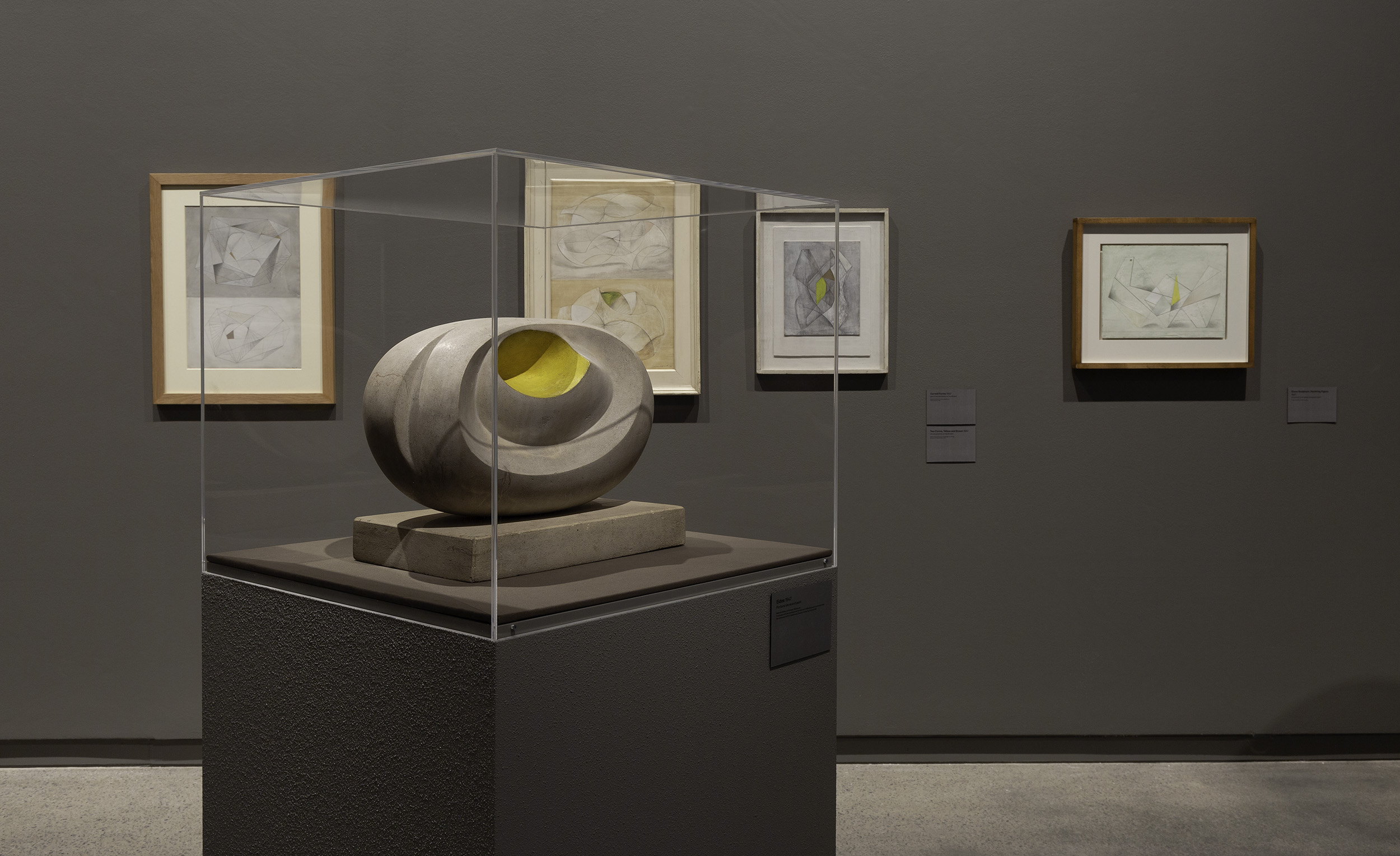 In a grey-white gallery, four framed abstract sketches hang on the wall behind a plinth holding a sculpture in a glass box. The sculpture is grey and a soft oval shape, with a piece scooped out of the middle and a green-painted hole left behind.