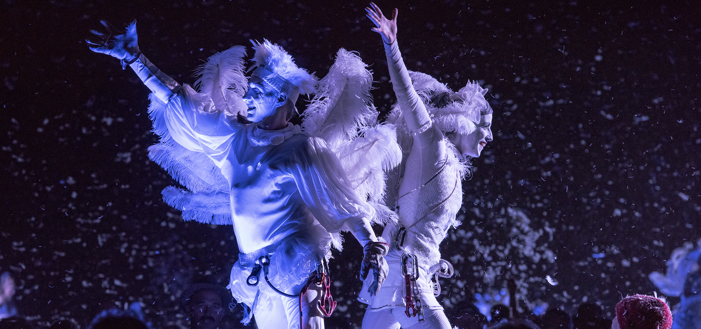 Two performers dressed in white costumes, their hands raised as feathers fall around them.