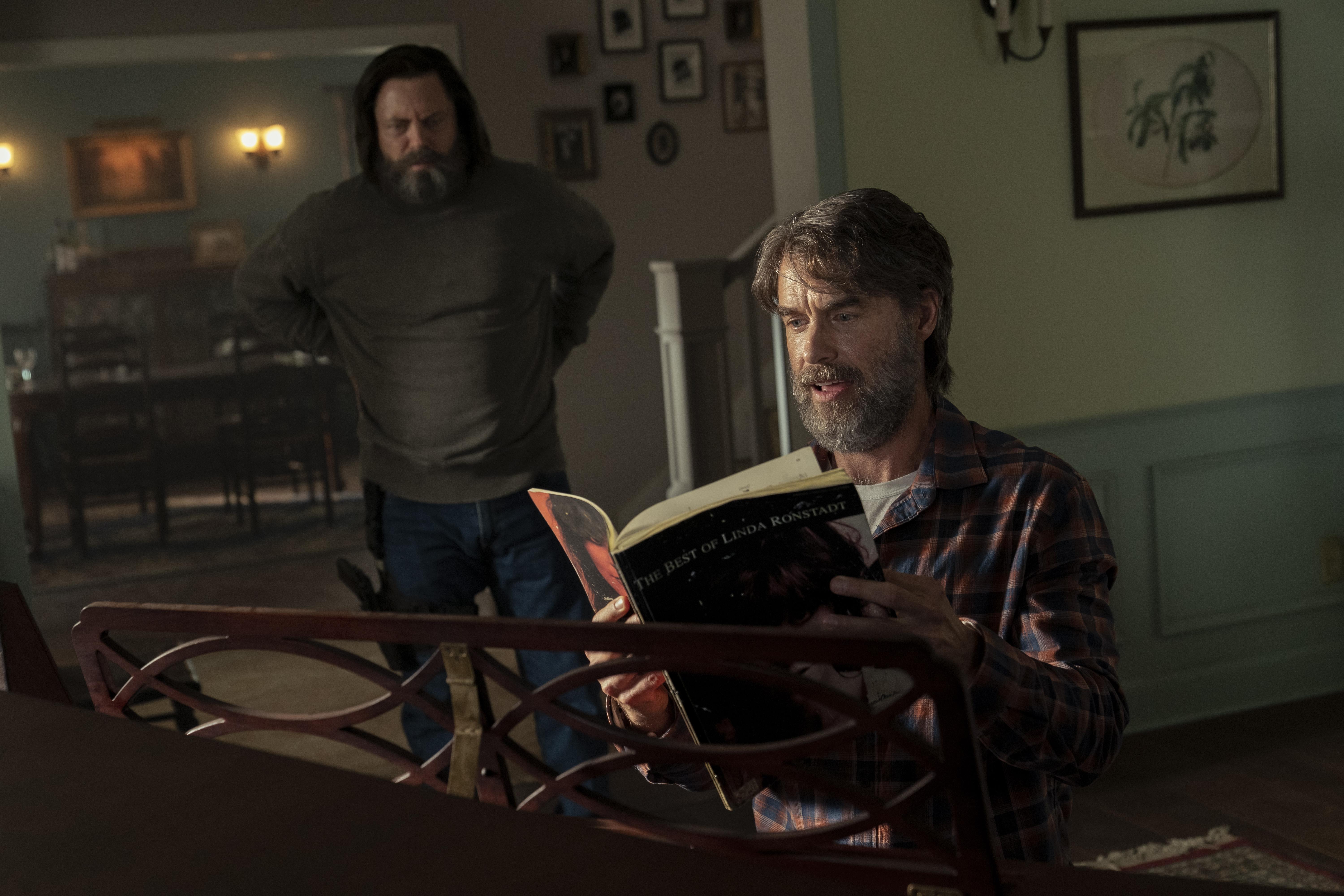 A middle-aged man wearing a greybeard and flannel shirt sits by a piano, looking through the pages of a Linda Ronstadt songbook. A man in similar clothing watches him from behind, gun strapped to his hip.