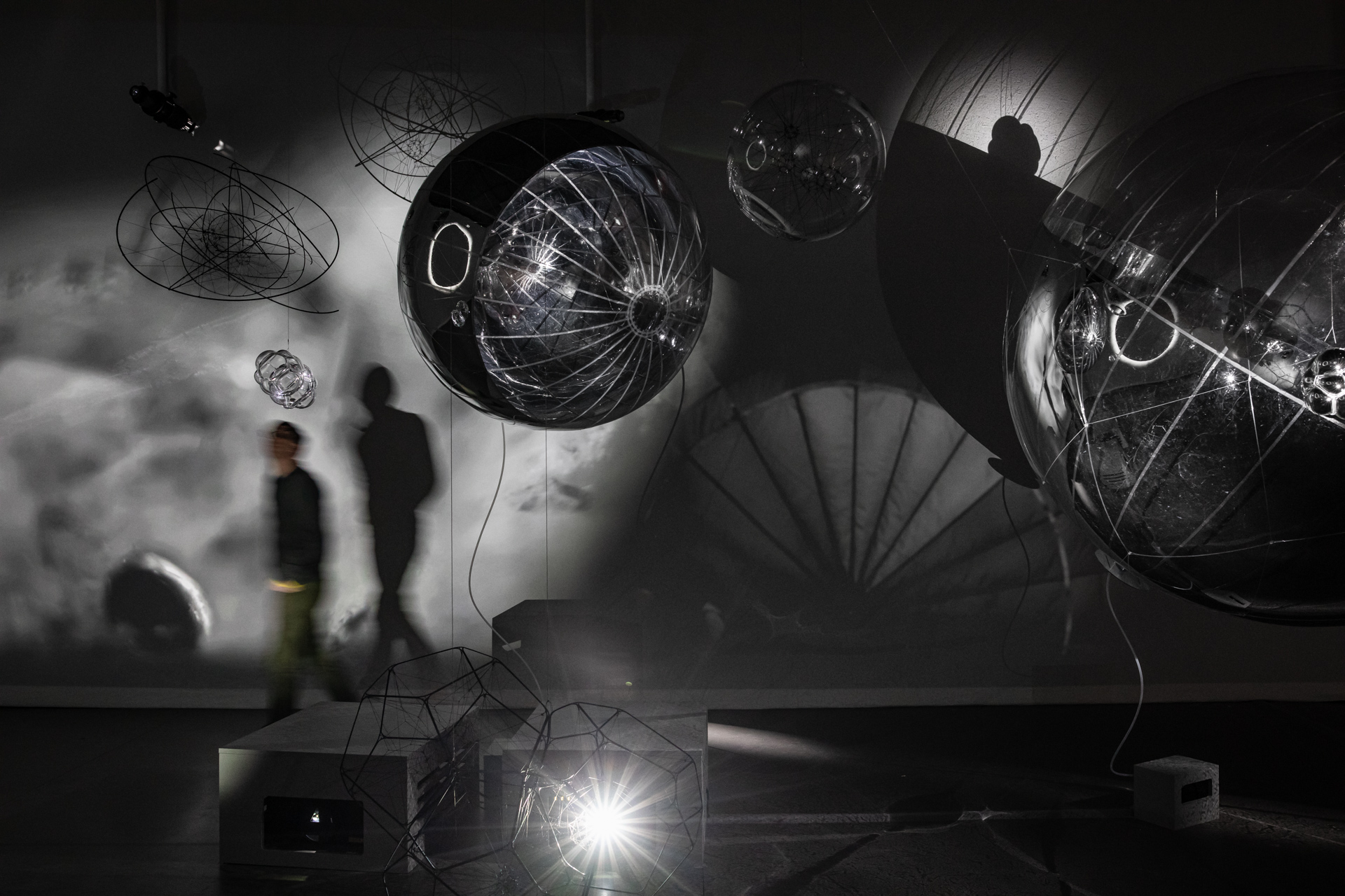A darkened gallery space in mostly black colours showcases fantastical ball-shaped metal sculptures with wires and hanging threads, with a person walking past blurred