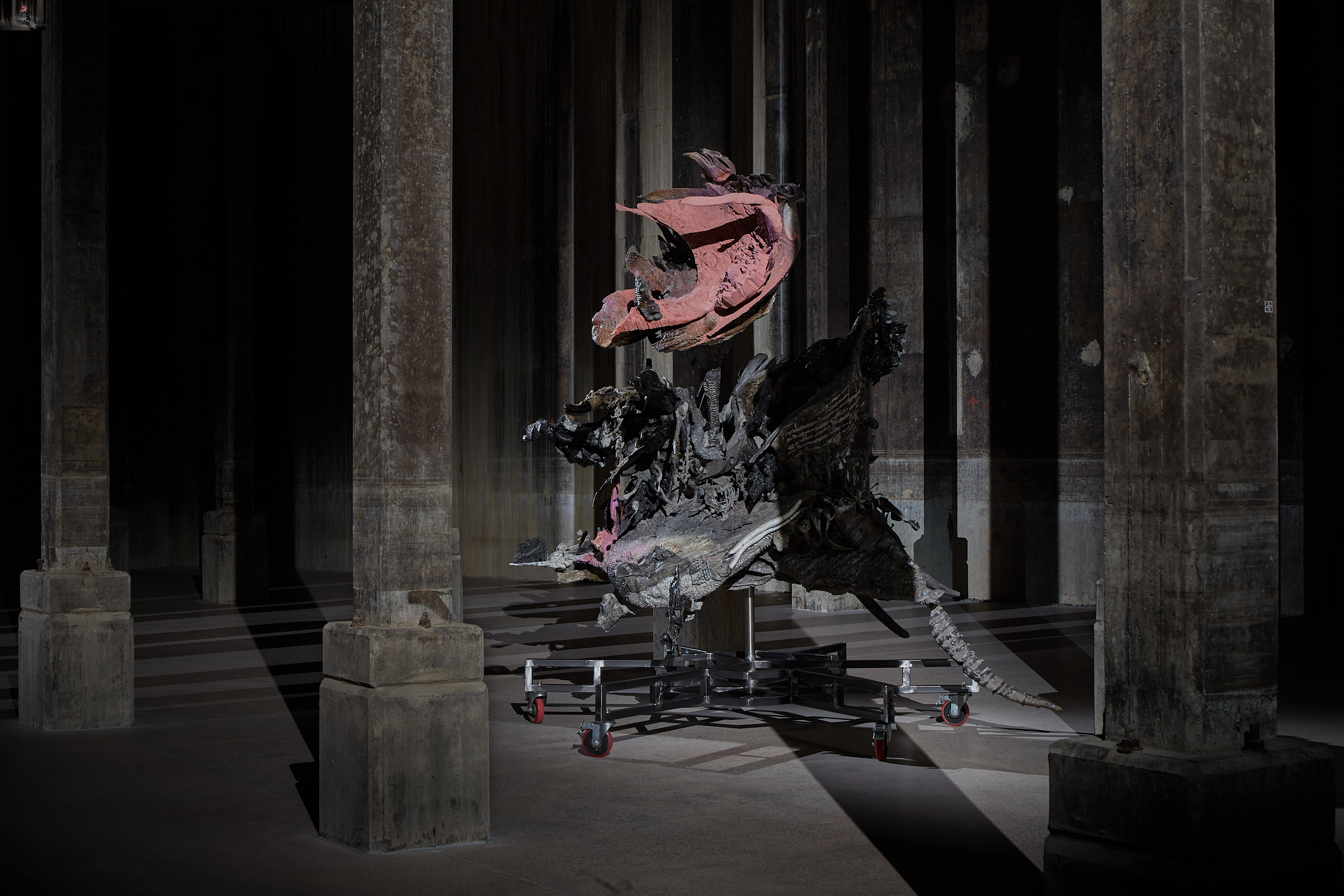 A grey and red faded dinosaur-like sculpture, with varying textures that appears prehistoric is mounted on a metal stand. It appears to be in a grey concrete hall, with the image quite dark and moody.