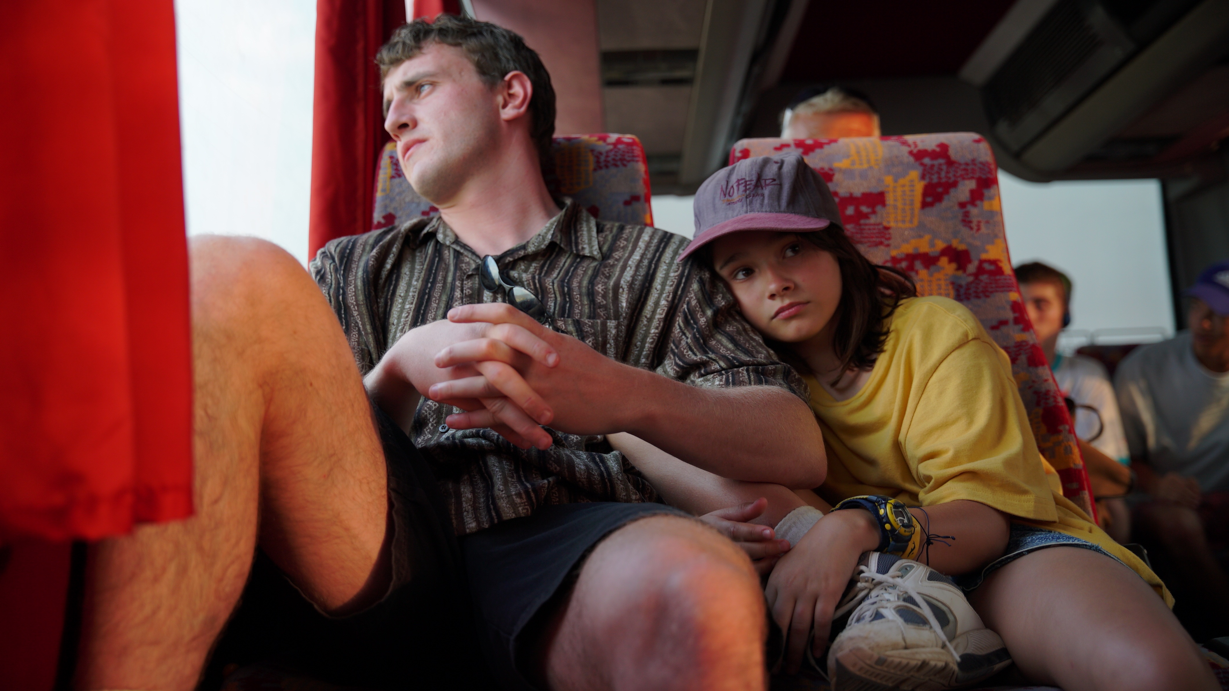 A man in shorts gazes out the bus window as a young girl in a baseball cap rests her head on his shoulder, looking toward the front of the bus. Her knee is raised up by the man's side.