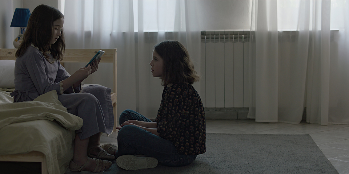 A young girl sits on her bed and is preoccupied with an iPad in her hand while another girl sits on the floor and watches her.