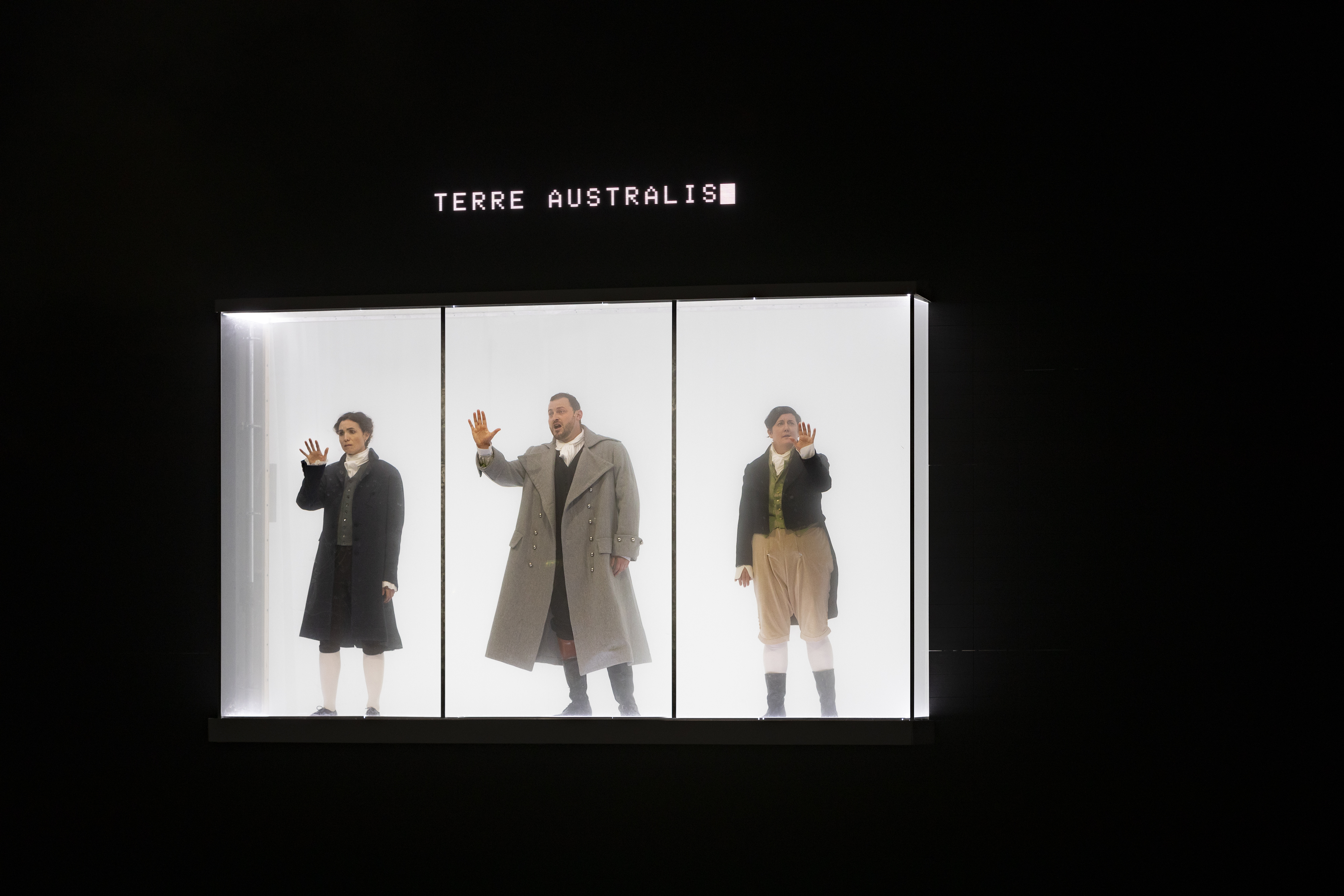 A white glass box surrounded by darkness holds three people wearing 18th century garb, they are all singing and holding one hand up to the glass. Above the box is white text reading "Terra Australis".