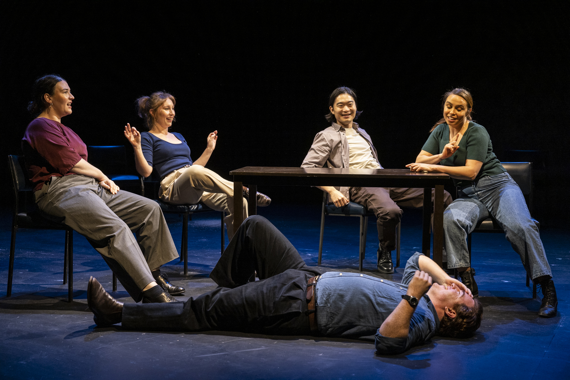 A group of performers sit in chairs around a desk. They're all looking down at another performer, who is flat on their back.