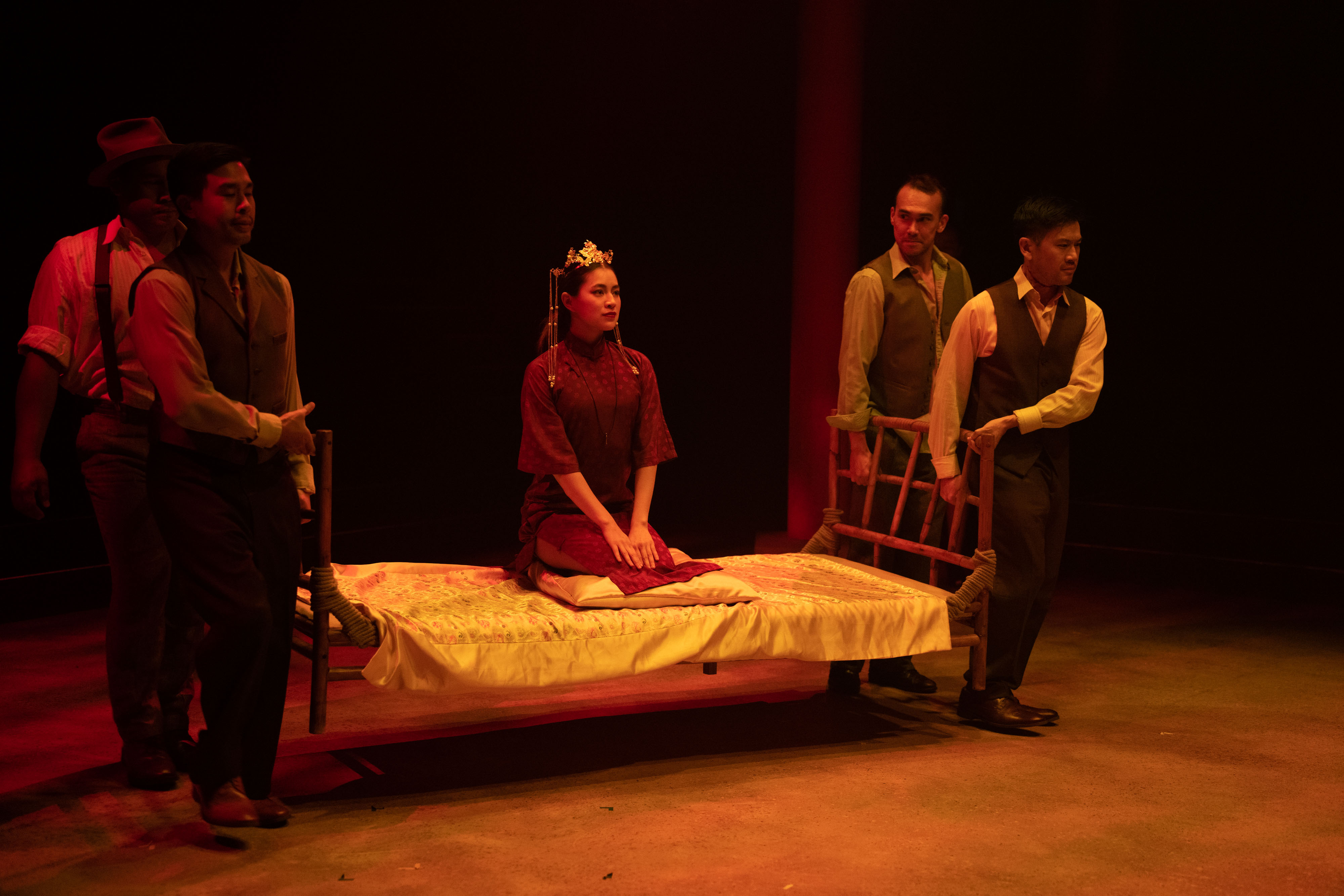 Four performers carry a bed with a woman wearing a crown sitting on top.