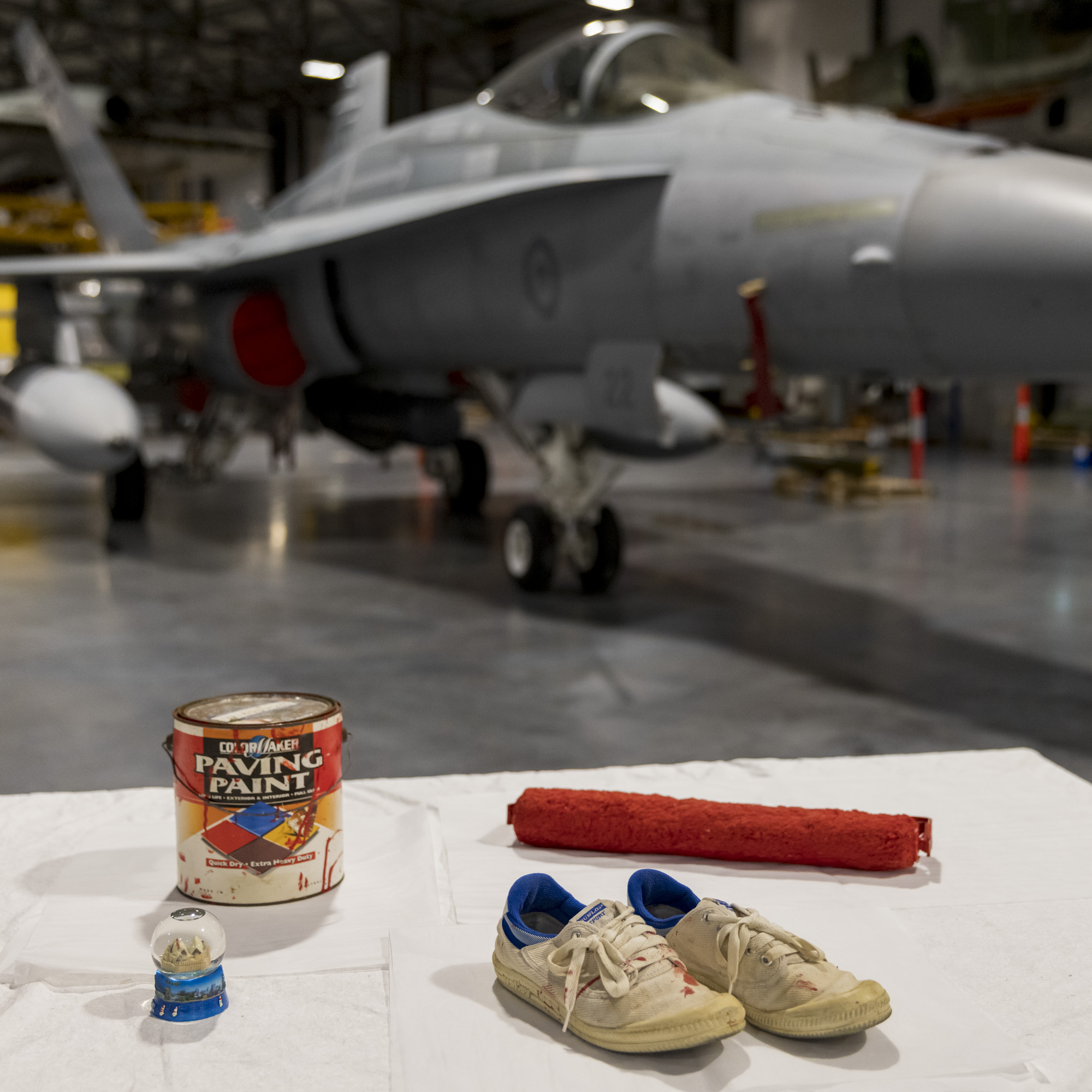 A tin of paint, shoes and other items displayed on a white table with a Hornet jet in the background.