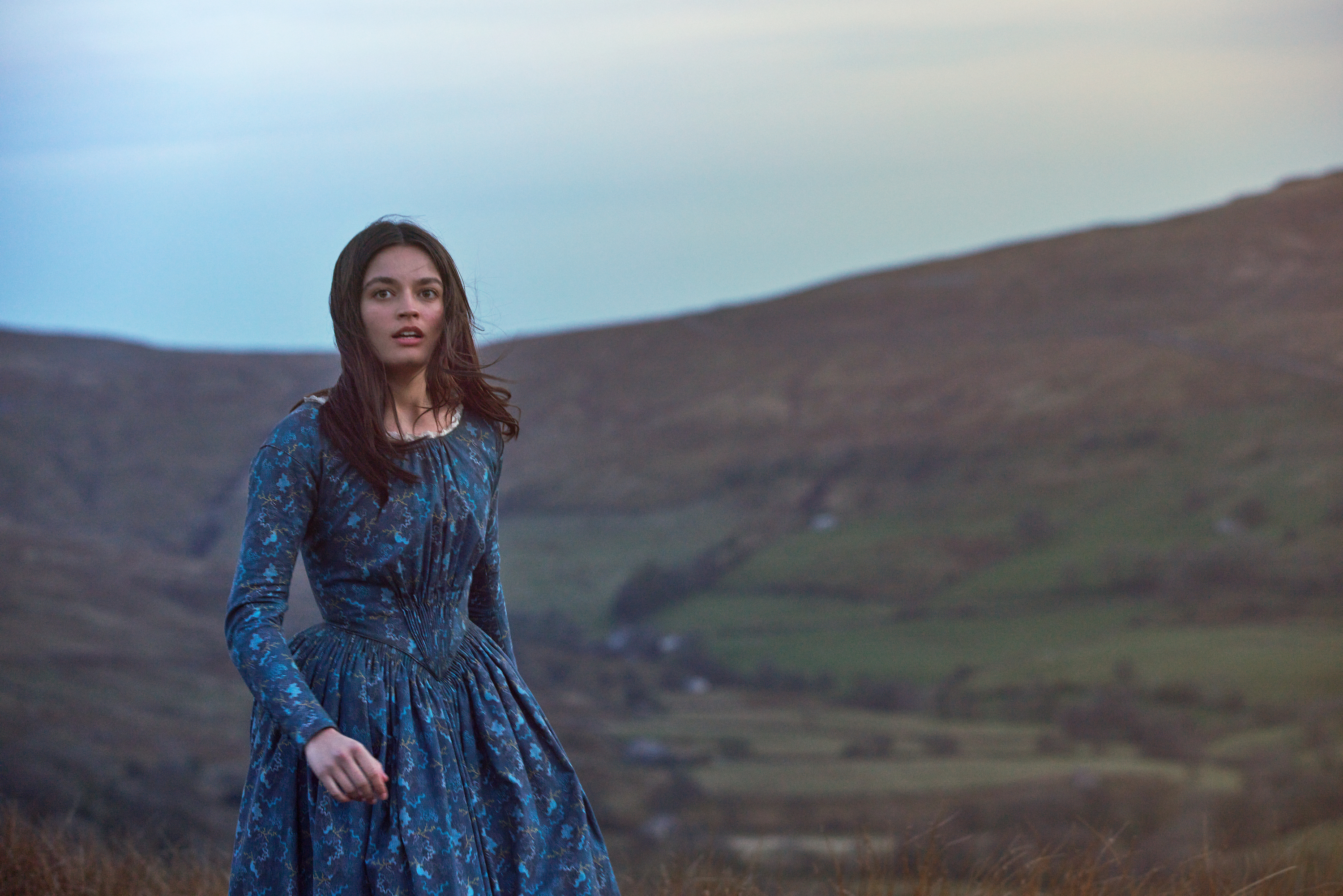A young Caucasian woman with pale skin and brunette hair wearing a patterned blue period dress appears windswept; she is somewhere outside on what looks to be an English moor